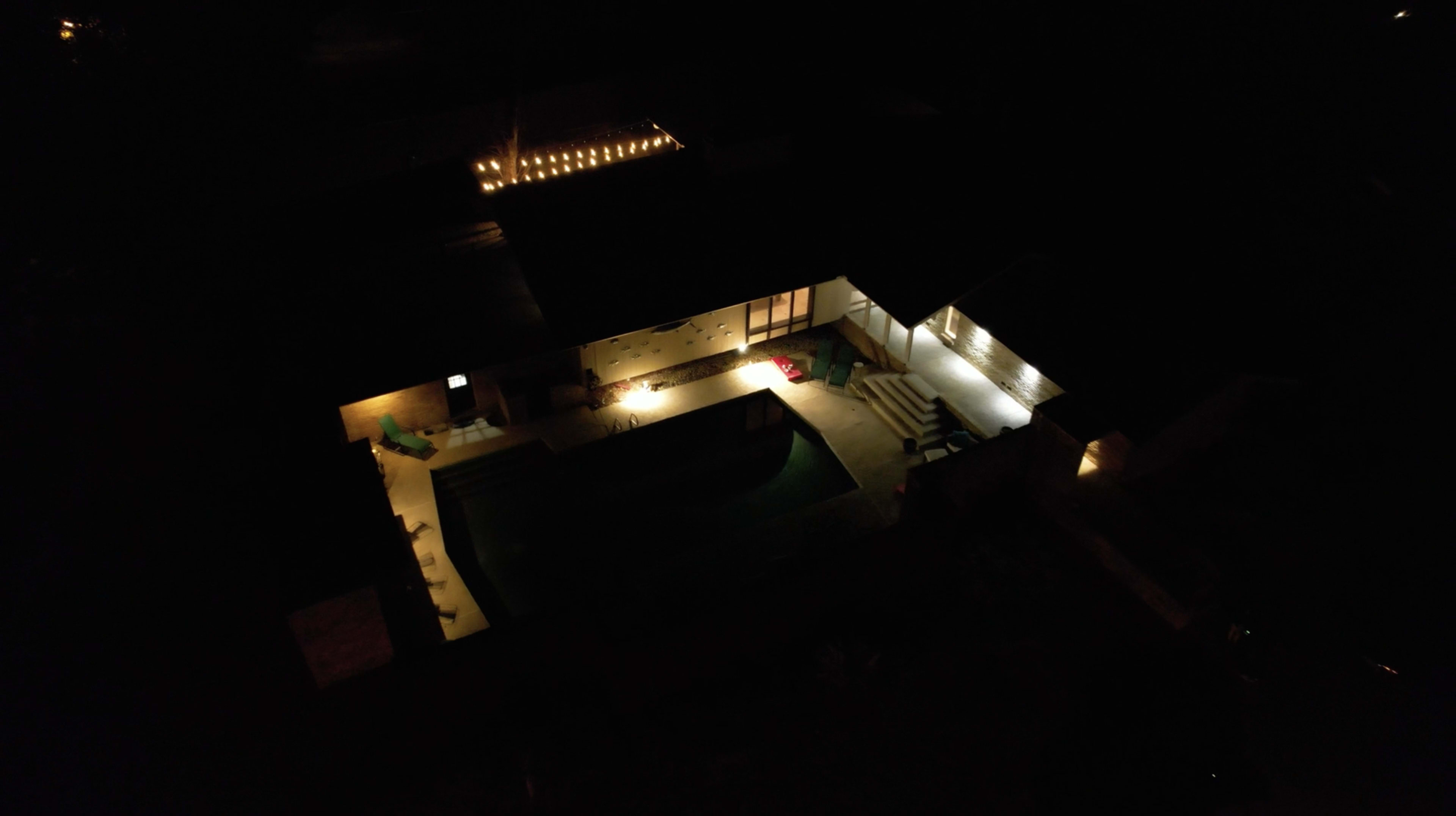 Aerial view of a house at night, illuminated by exterior lights around a swimming pool.
