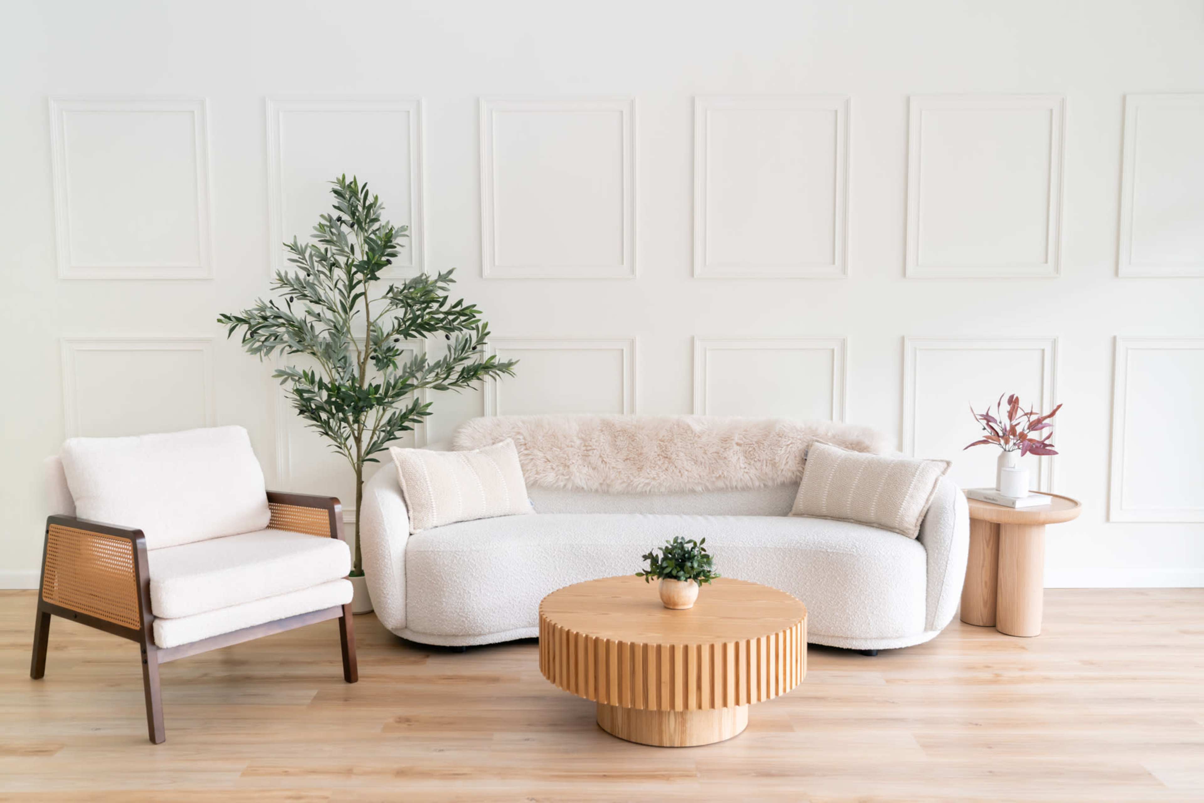 The image shows a modern living room setup featuring a white, curved sofa, a wooden coffee table with vertical slats, a light armchair, and decorative plants against a plain wall with paneling.