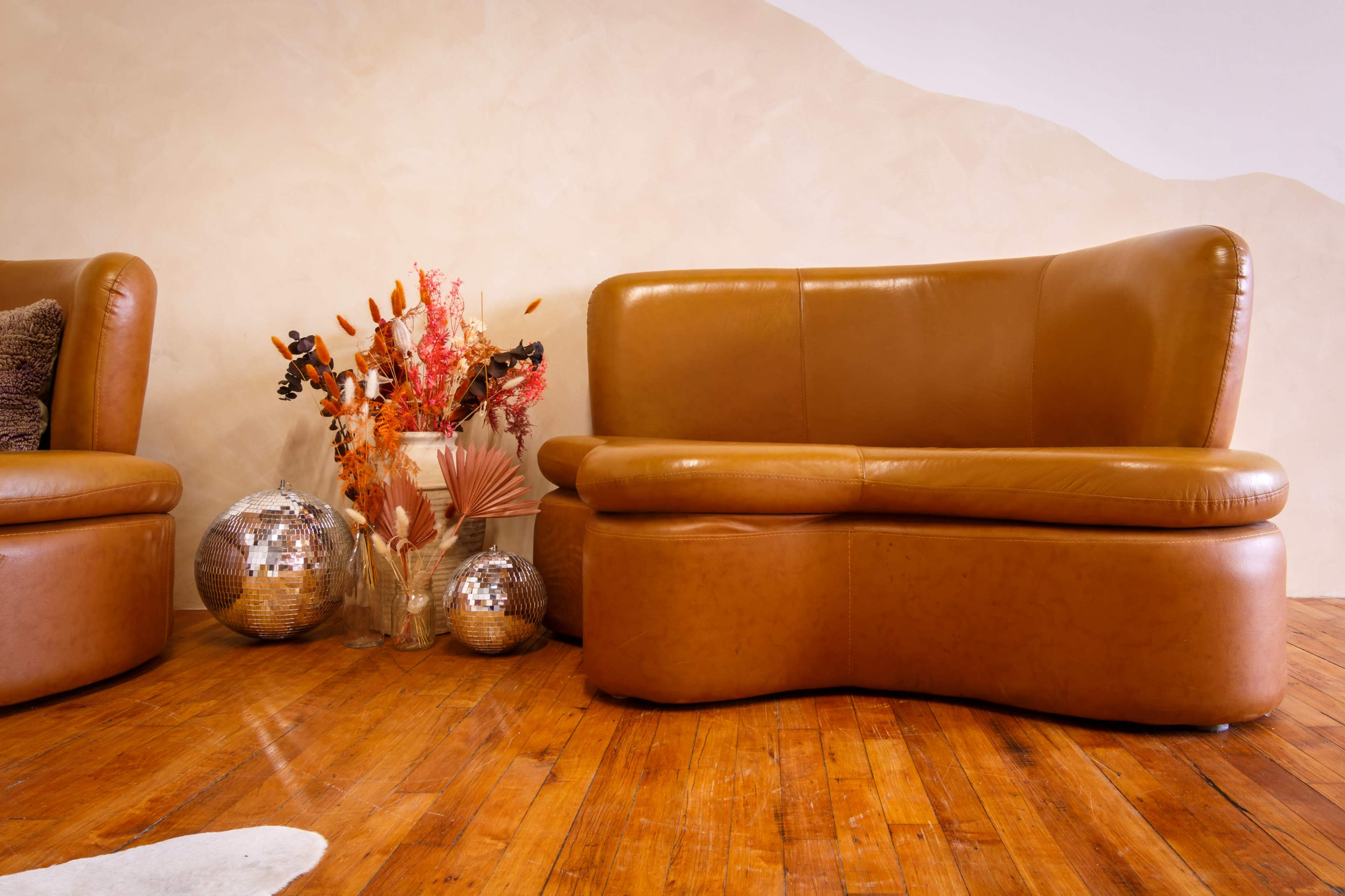 A curved brown leather sofa sits beside a vase of dried flowers and two disco balls on wooden flooring.