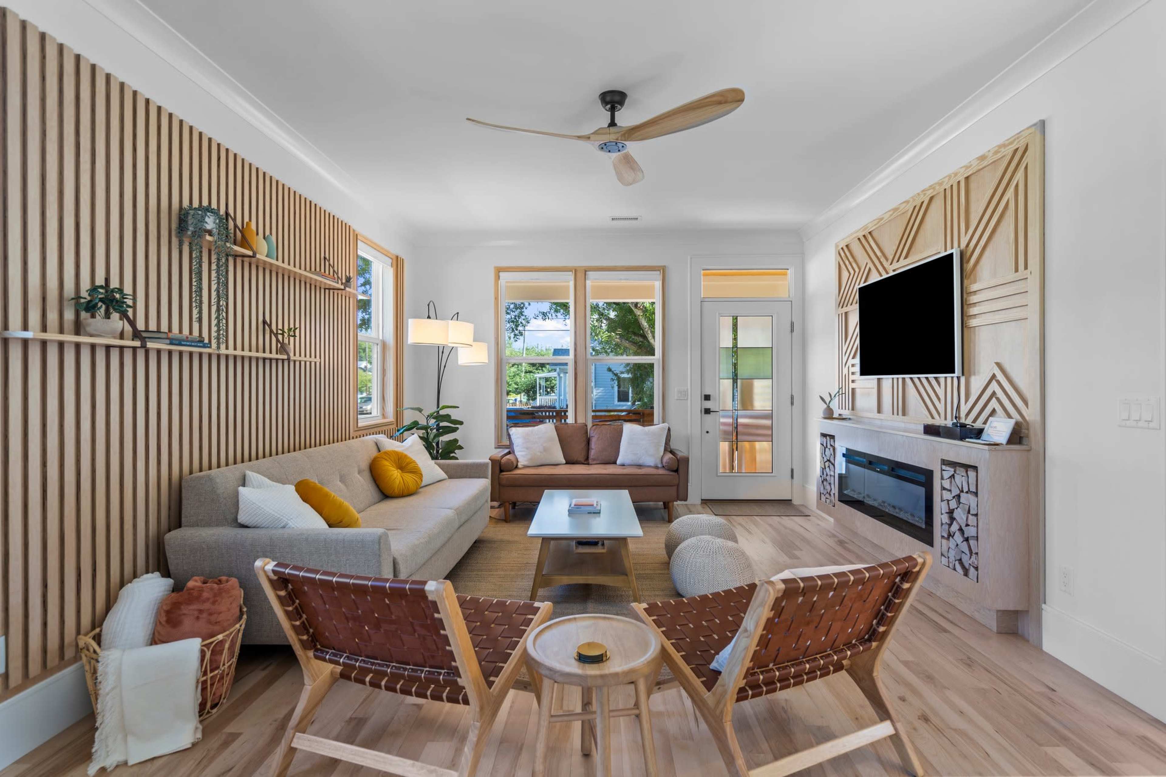 The living room features a light-colored sofa, two brown chairs, and a wooden accent wall with large windows and a door leading outside.