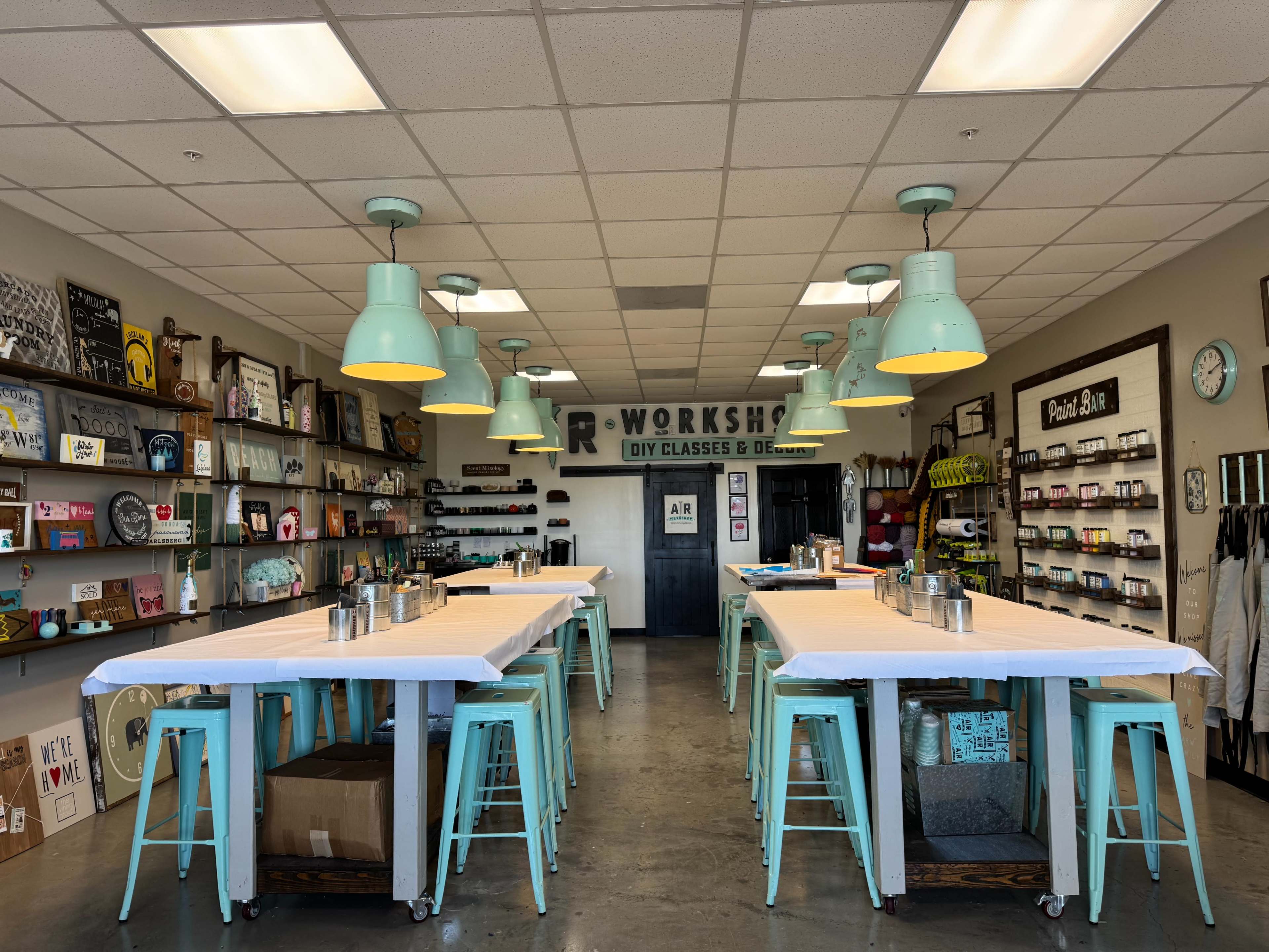 The image shows a brightly lit workshop space with multiple work tables, teal stools, and shelves lined with art supplies and decor items.