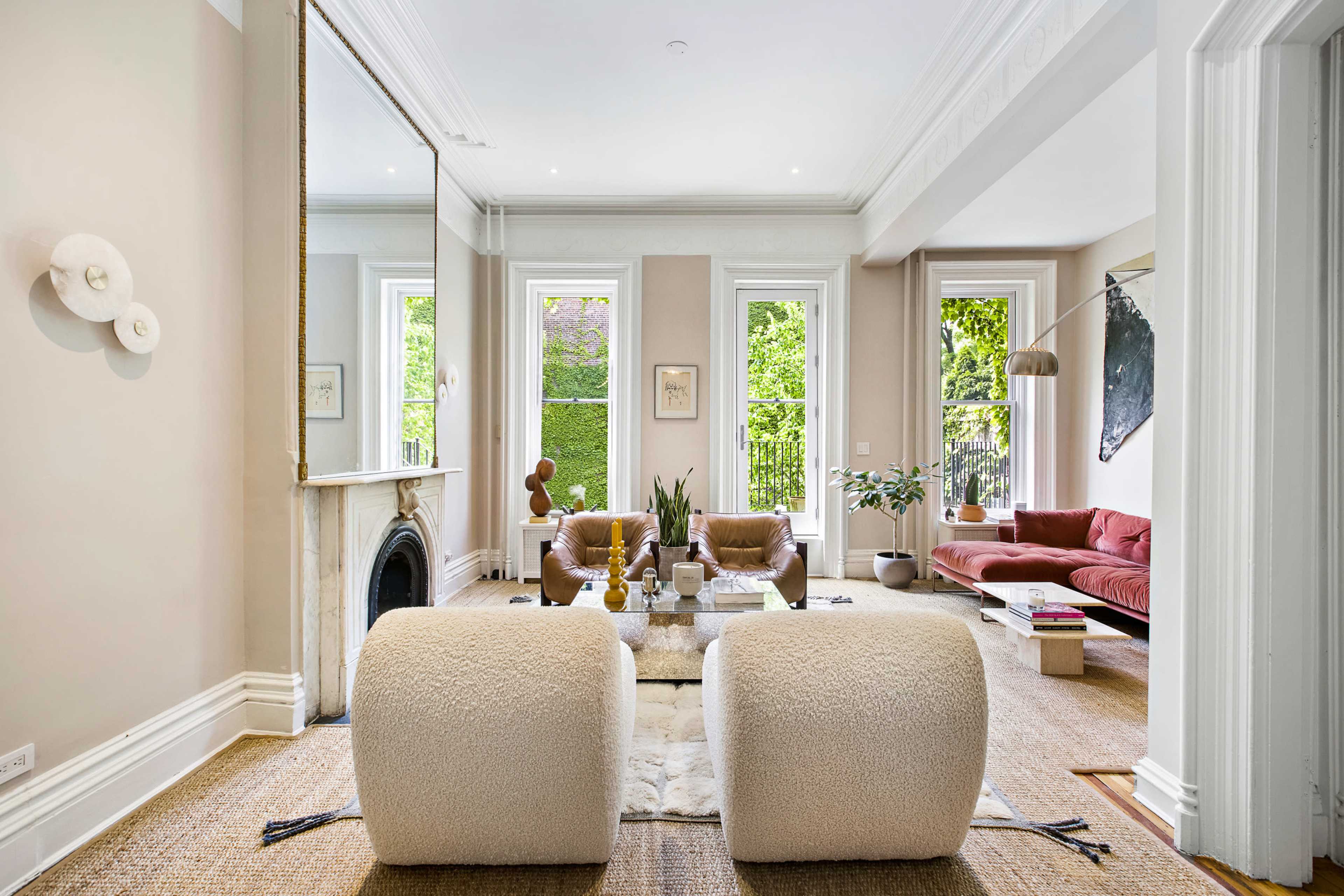 A bright living room features a pink sofa, a fireplace, and large windows with greenery visible outside.