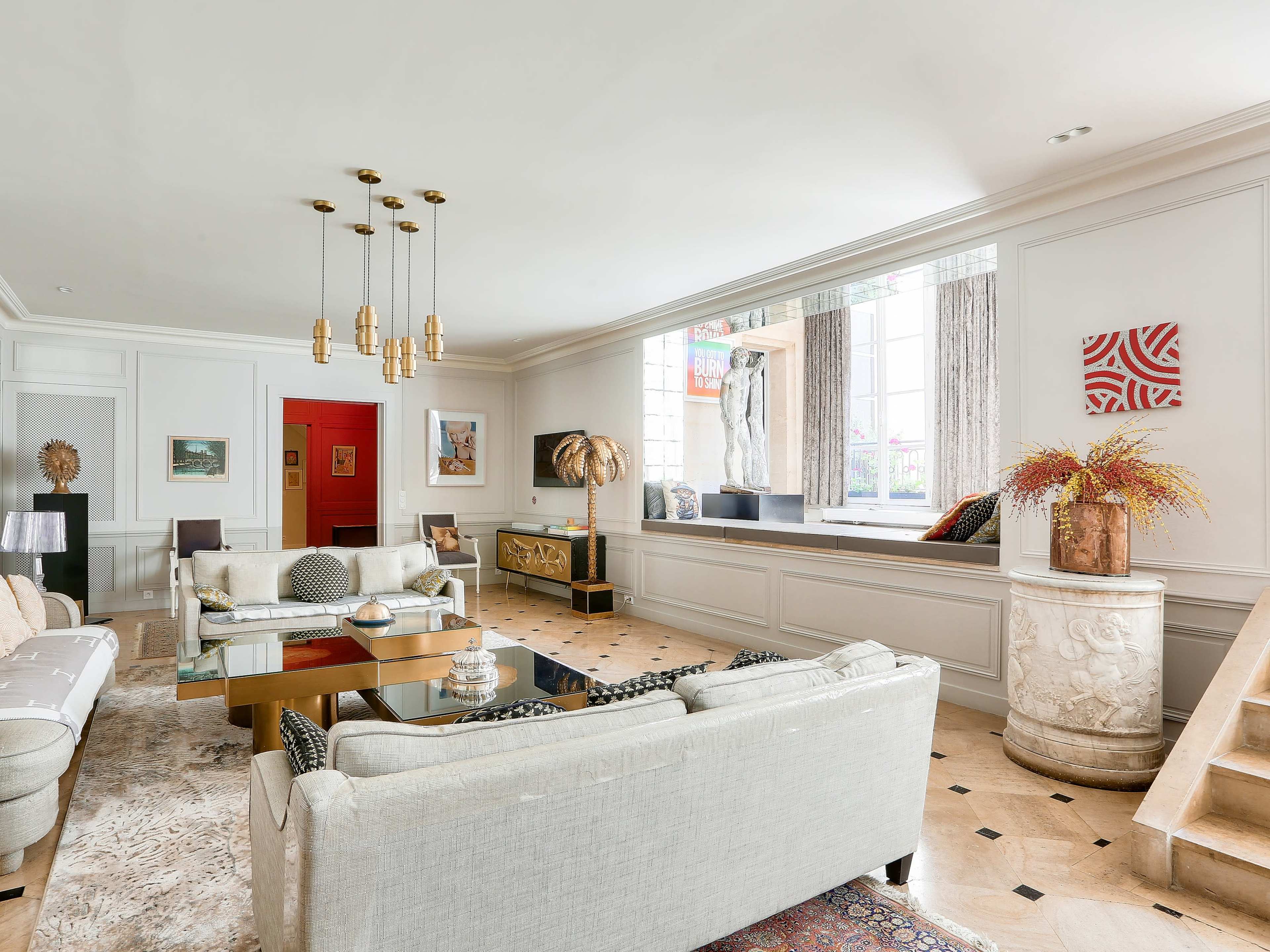 The image shows a spacious, well-lit living room with a seating area arranged around a glass coffee table, featuring decorative artwork and plants.