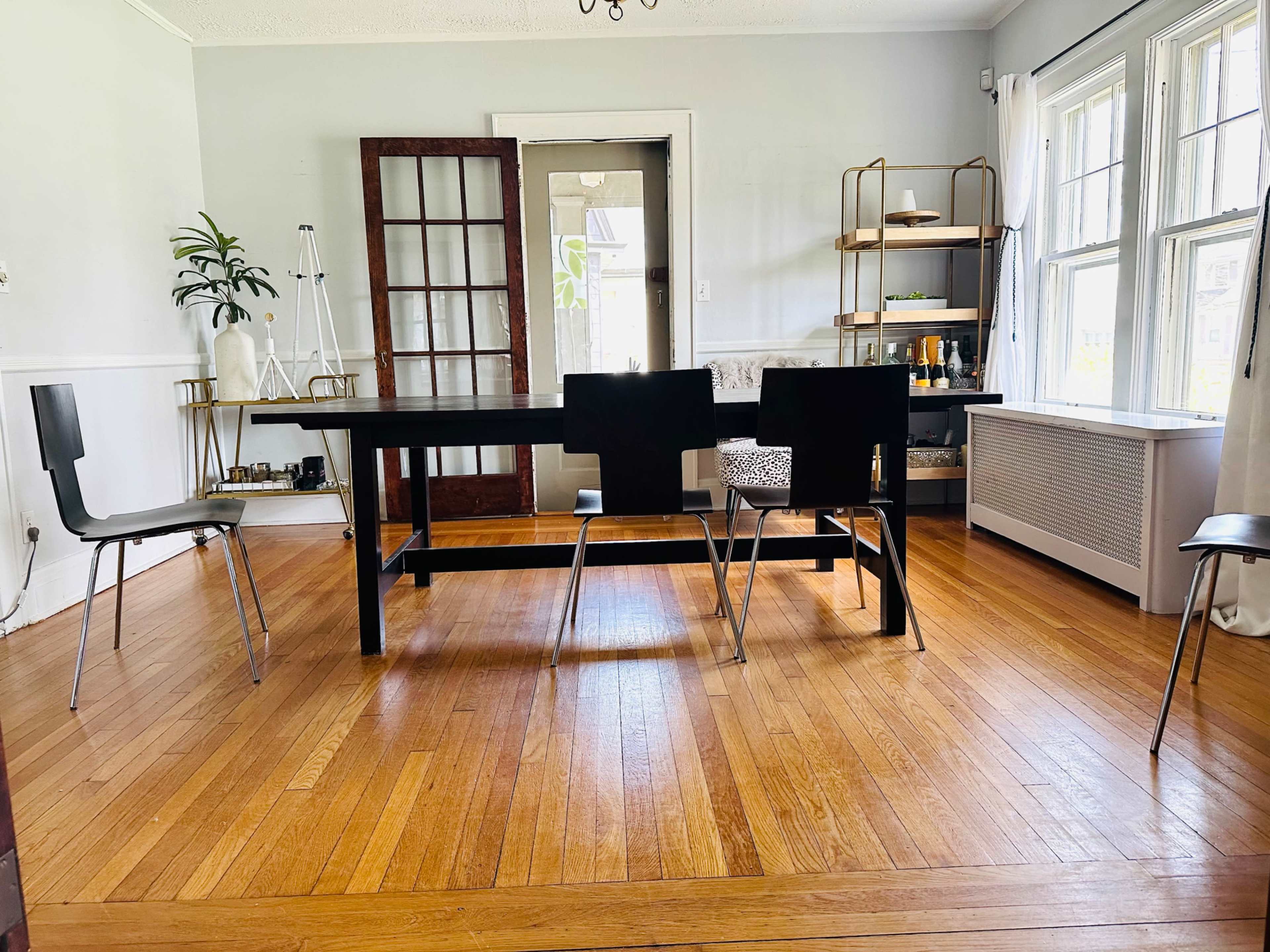 A spacious dining area features a large black table surrounded by several chairs on a hardwood floor, with plants and shelving visible in the background.