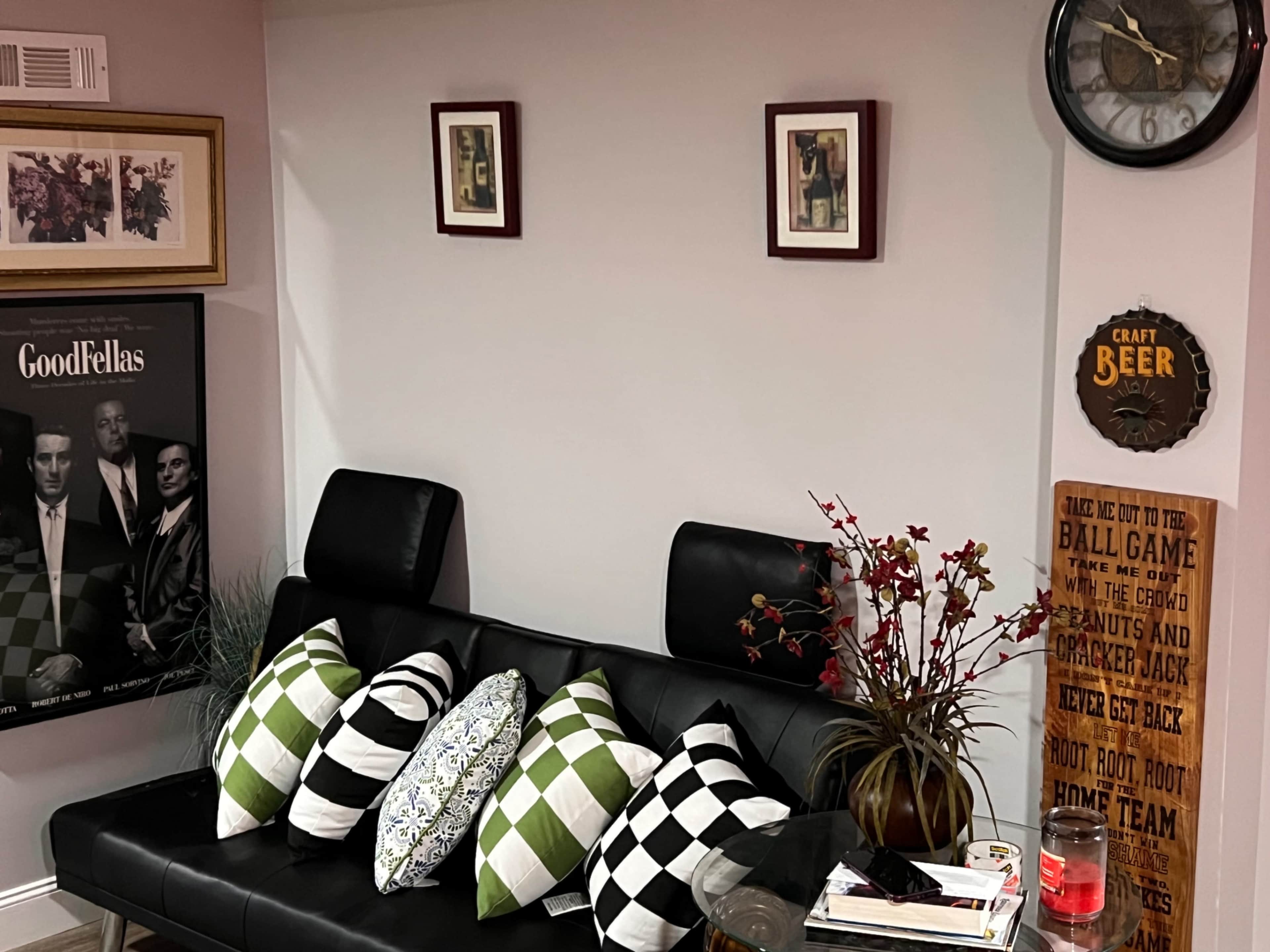 The scene features a black sofa with decorative pillows, a wall adorned with framed artwork, and a clock, all set in a cozy room with a sign reading "CRAFT BEER" and a small table with books and a candle.