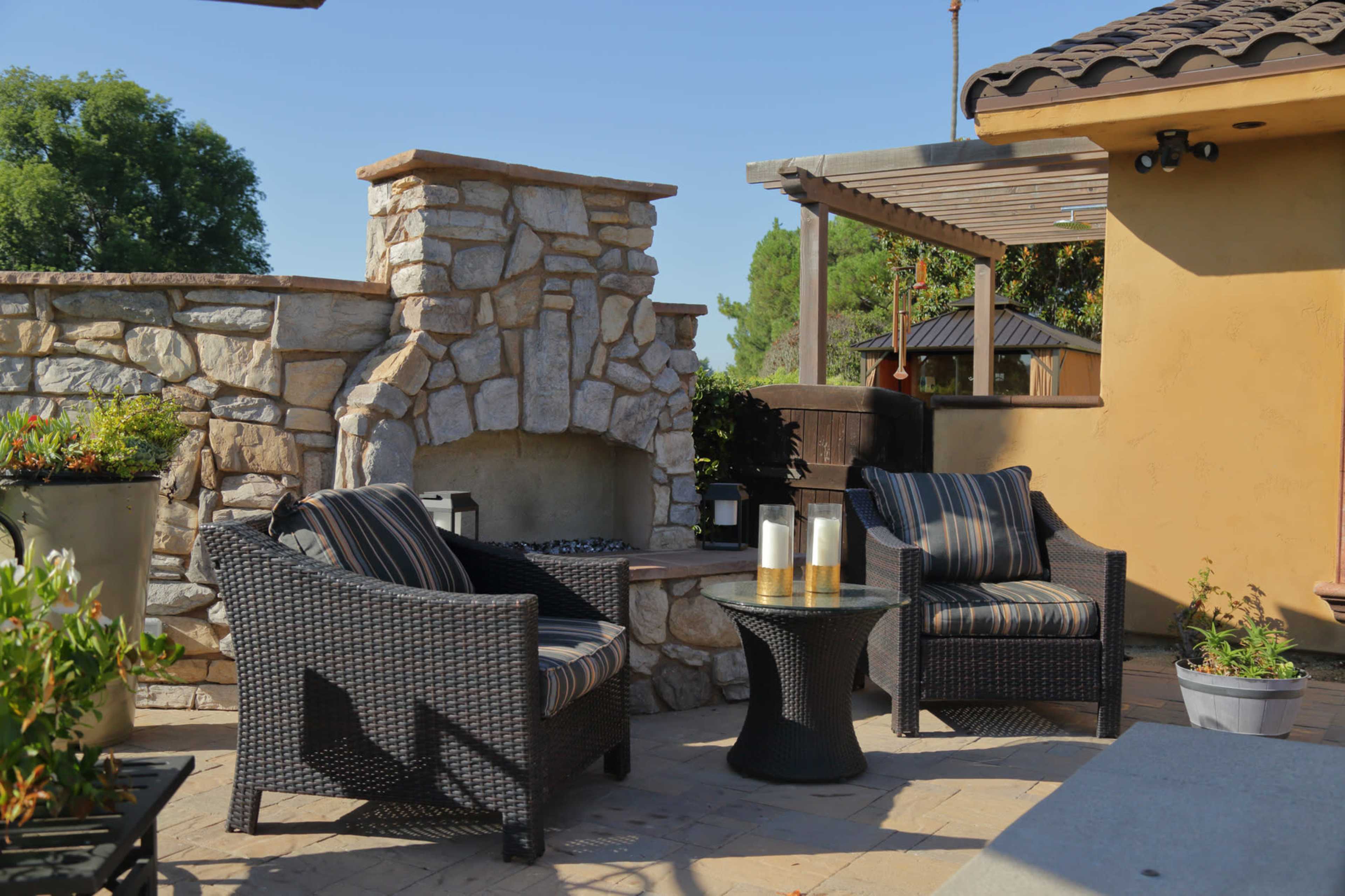 A patio features two wicker chairs with striped cushions positioned near a stone fireplace.