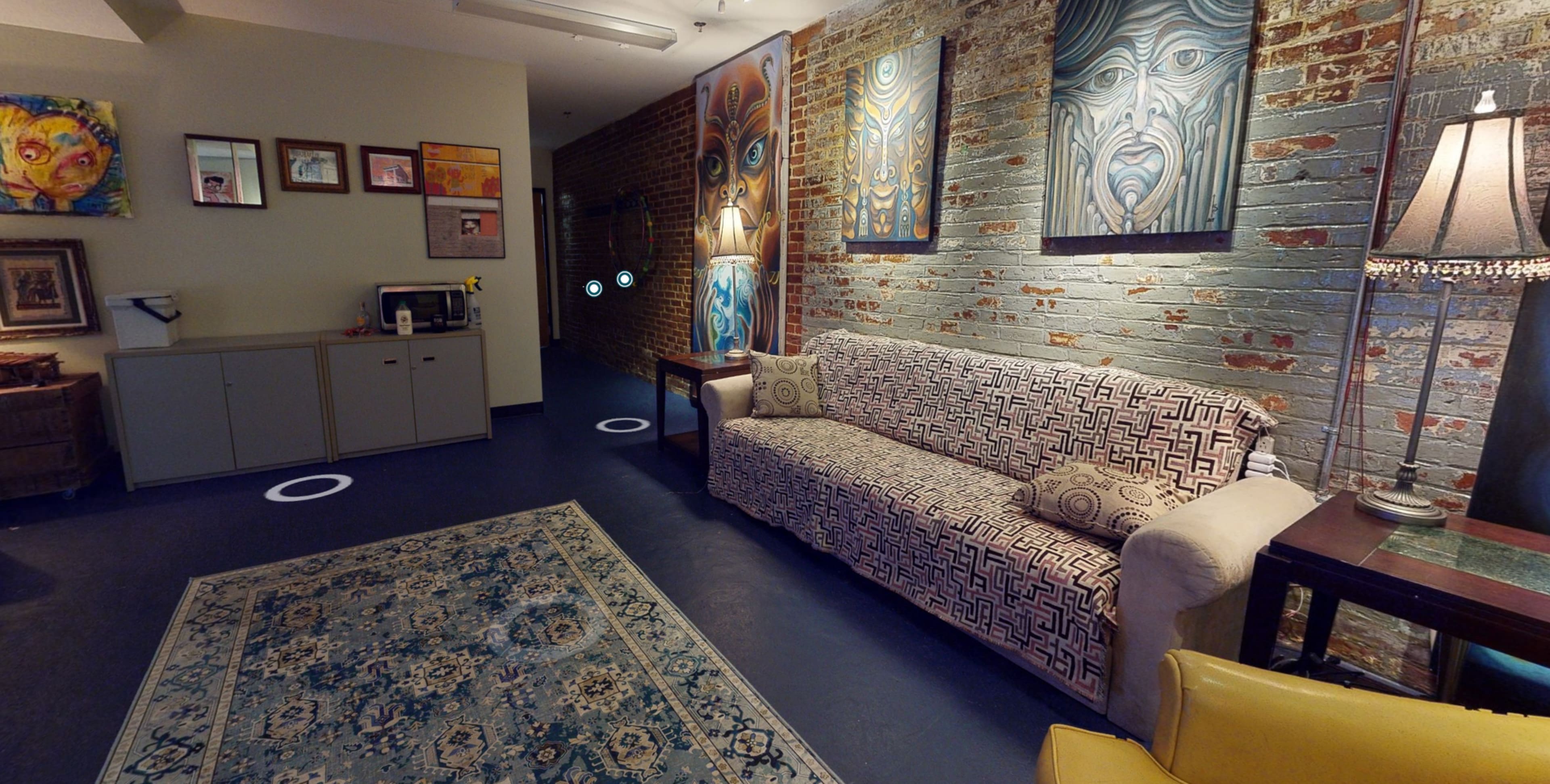 The image shows a neatly arranged room with a patterned sofa, framed artwork on the brick walls, and a decorative rug on the floor.