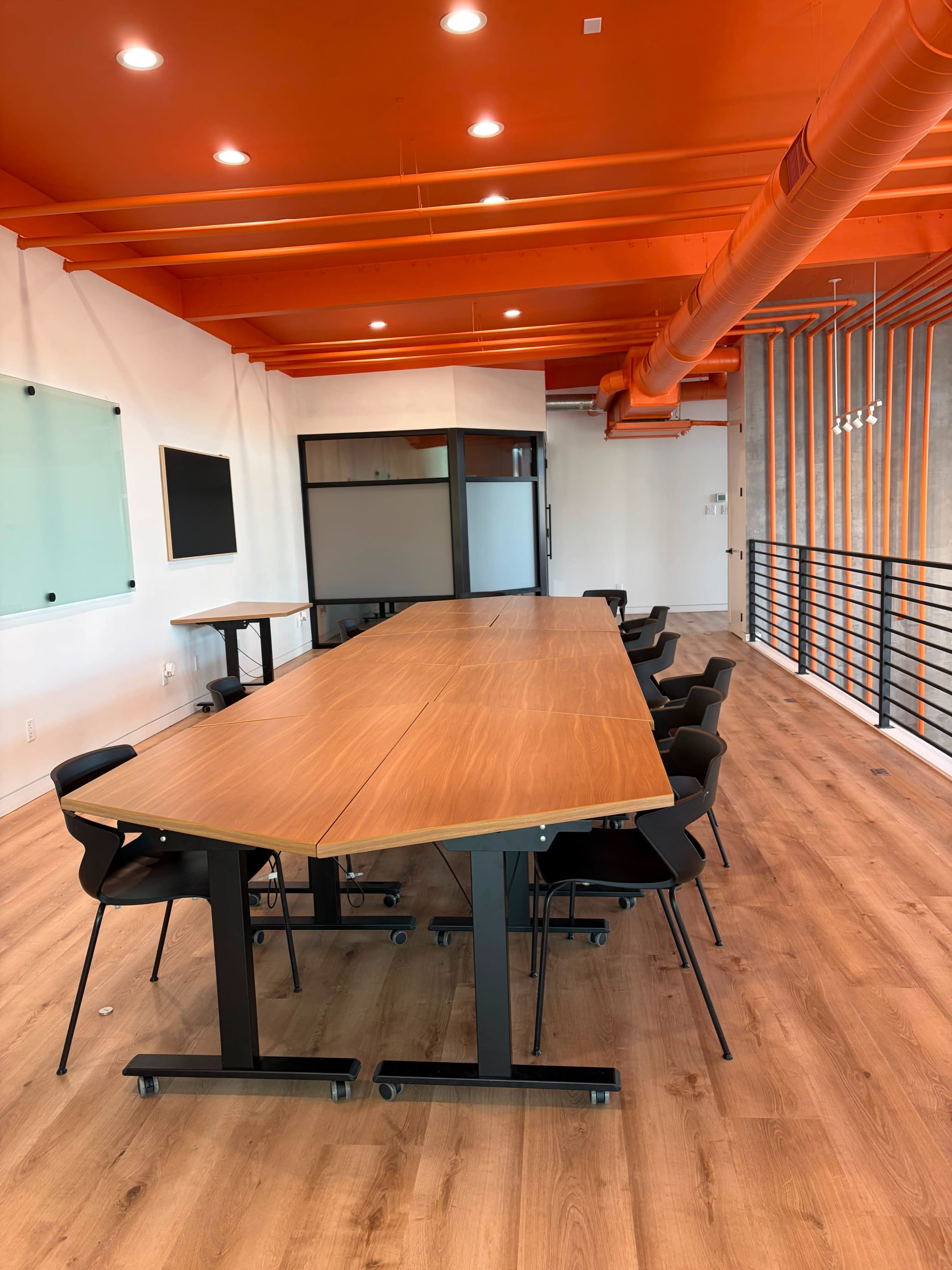 A long wooden conference table with black chairs is situated in a brightly lit meeting room with orange ceiling beams and walls.