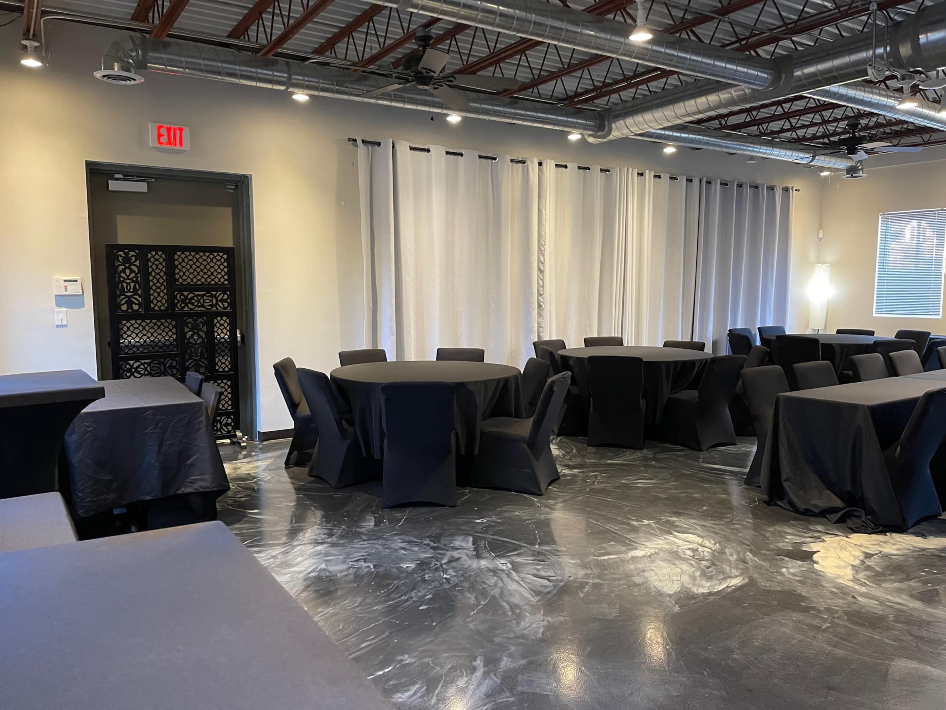 The image shows an event space with several black round tables and chairs arranged neatly in a well-lit area.