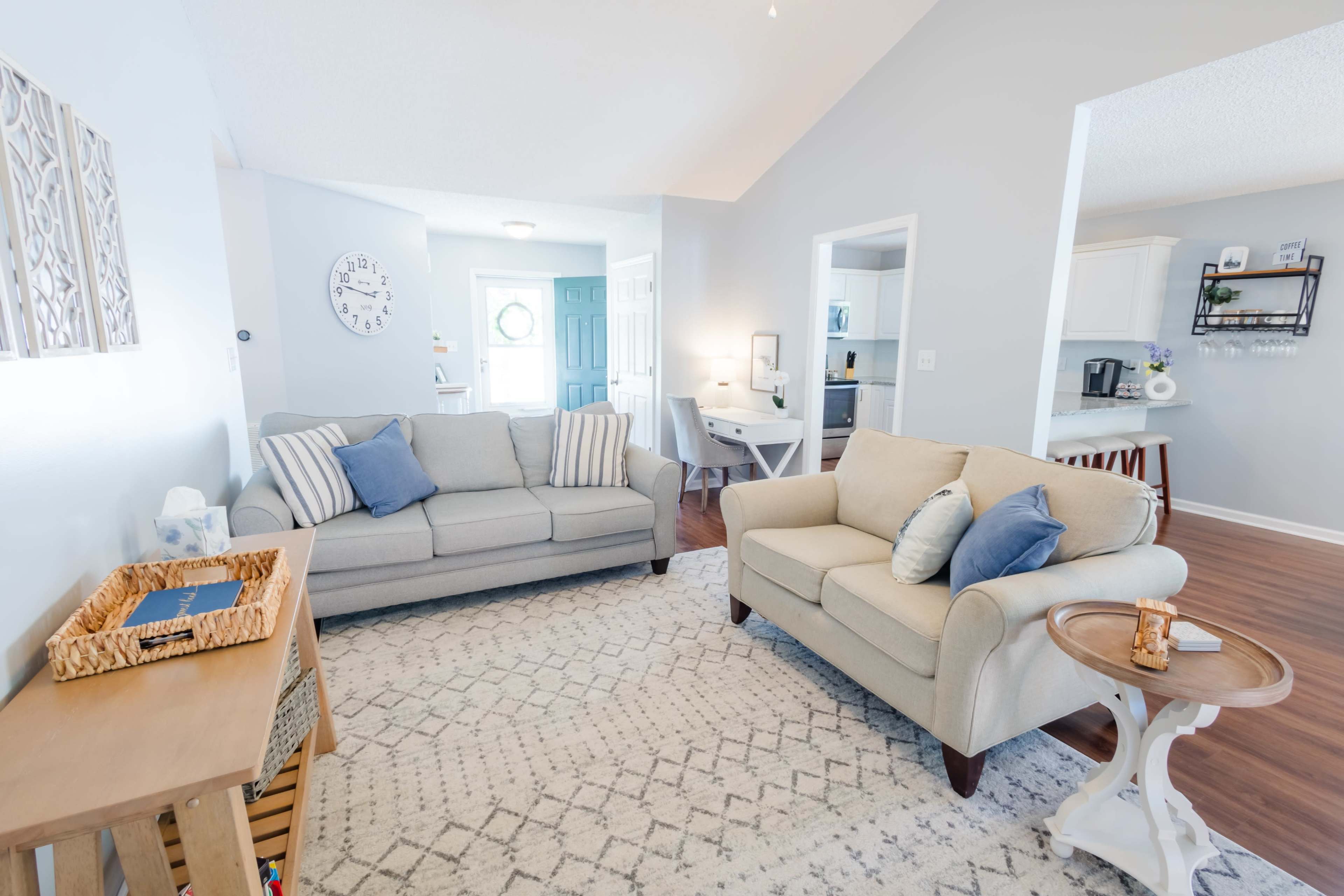 The living room features two sofas, a coffee table, and a light-colored rug, with a doorway leading to a kitchen area in the background.
