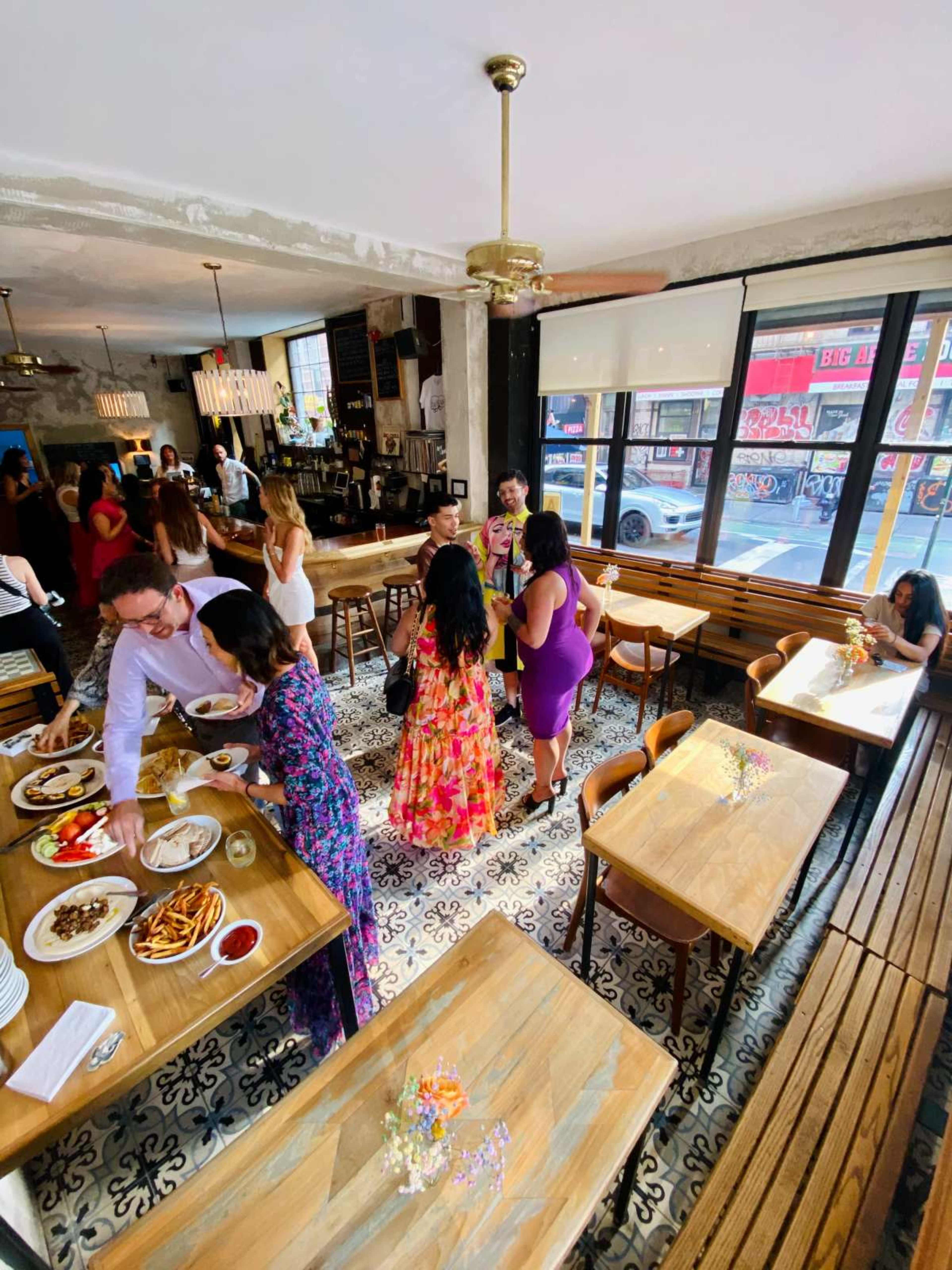 The image shows a bustling restaurant interior with diners socializing, tables filled with food, and a large window revealing a city street outside.