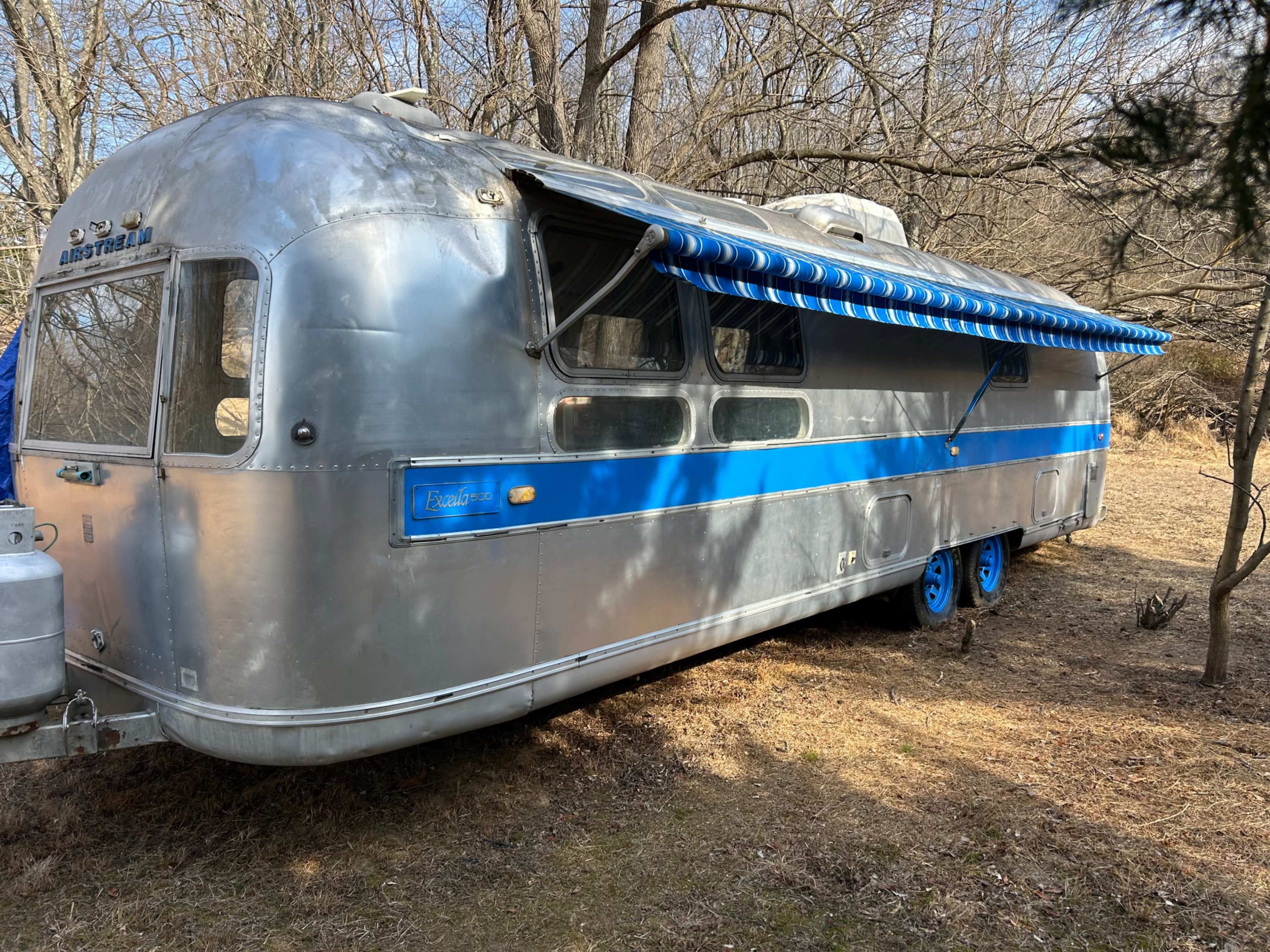 The image shows a vintage Airstream trailer parked in a wooded area with blue accents and a retractable awning.
