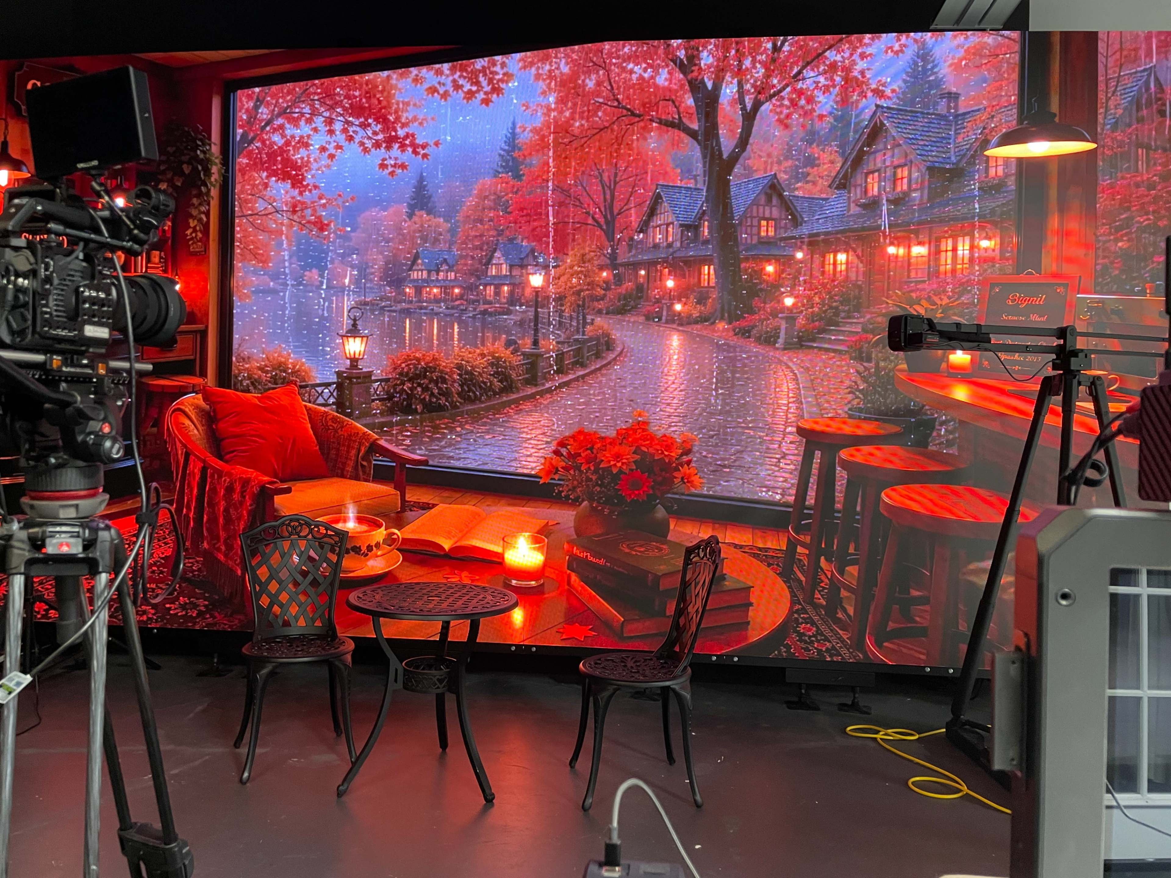 The scene features a cozy set with a large backdrop of a tranquil autumn landscape and rustic houses, complemented by two chairs and a table with candles in the foreground.