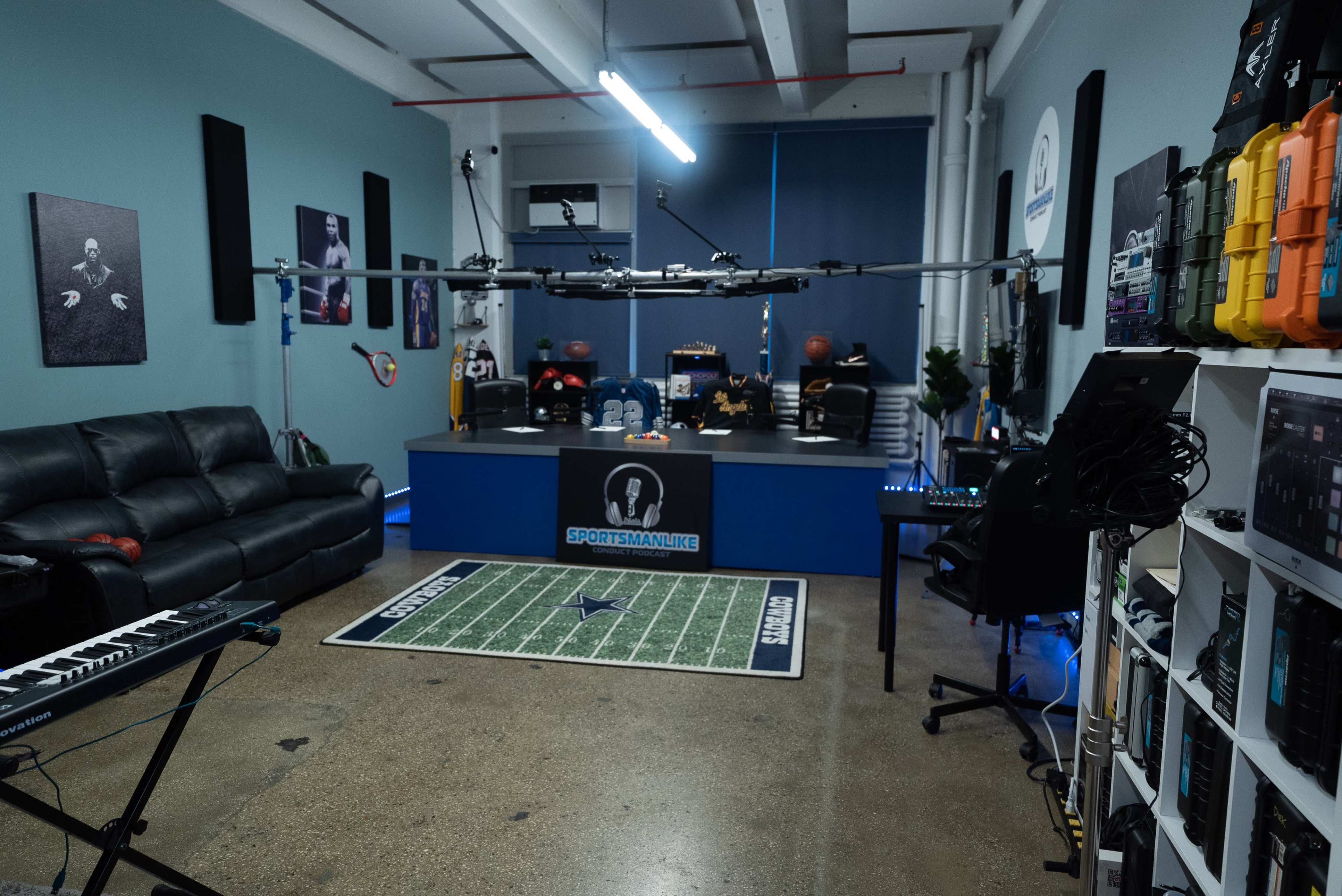 A modern studio setup featuring a black leather sofa, a desk with sports-themed decorations, and various equipment, including a keyboard and a shelf organized with tech gear.