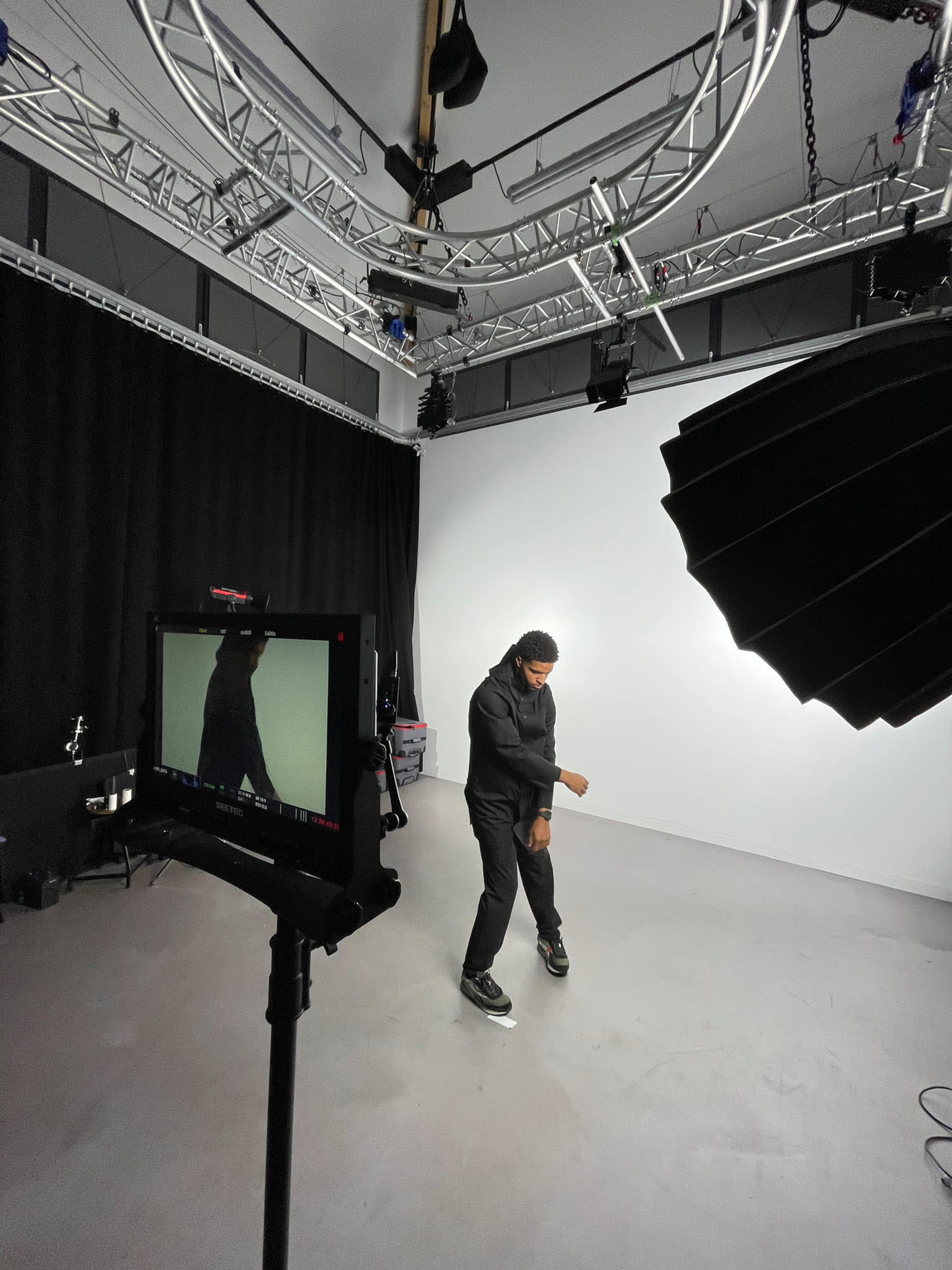 A person is dancing in a studio with bright lighting and a black backdrop, while a camera is positioned on a stand capturing the scene.
