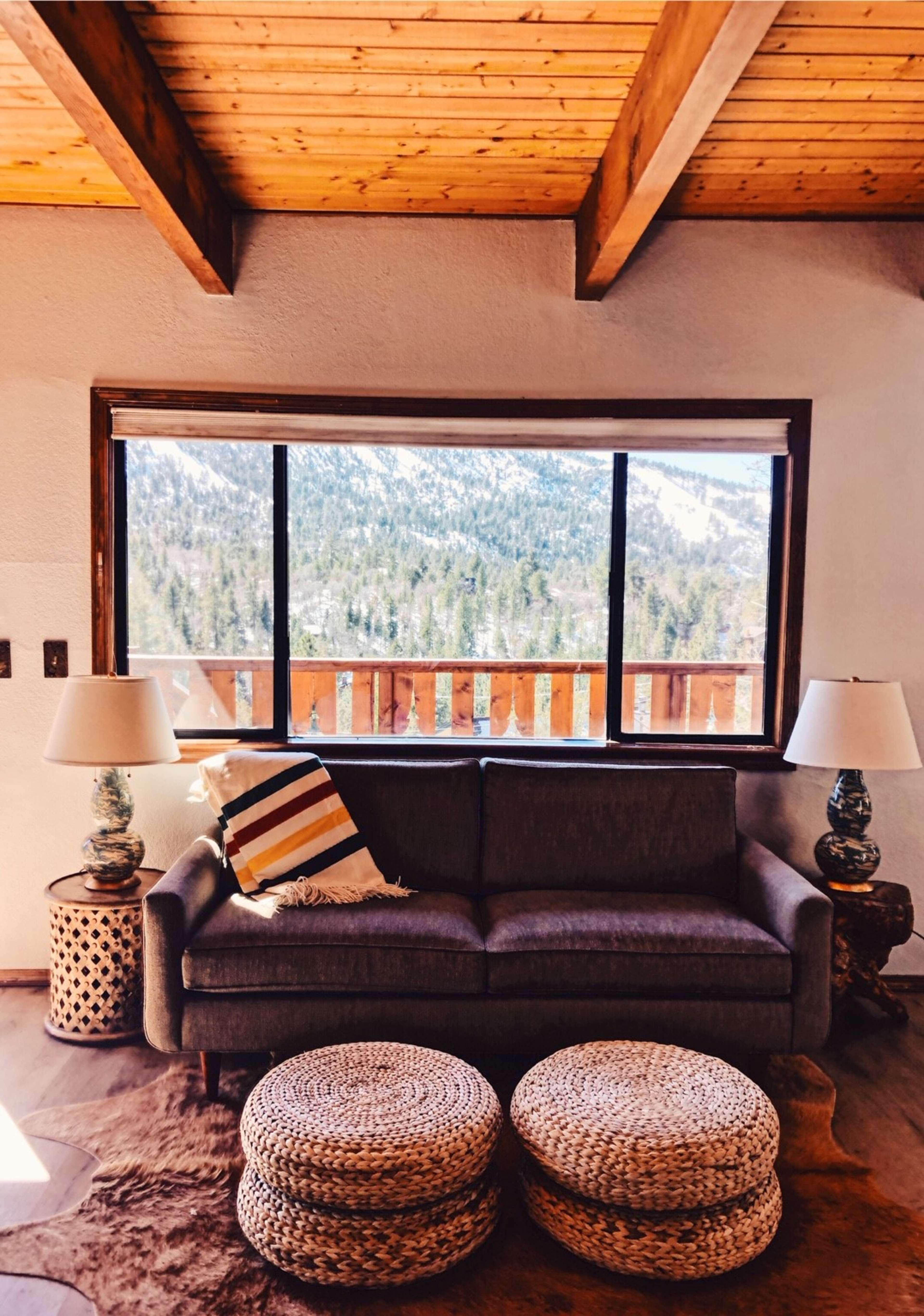 The image shows a living room with a gray couch, two woven ottomans, and large windows offering a view of a mountainous landscape.