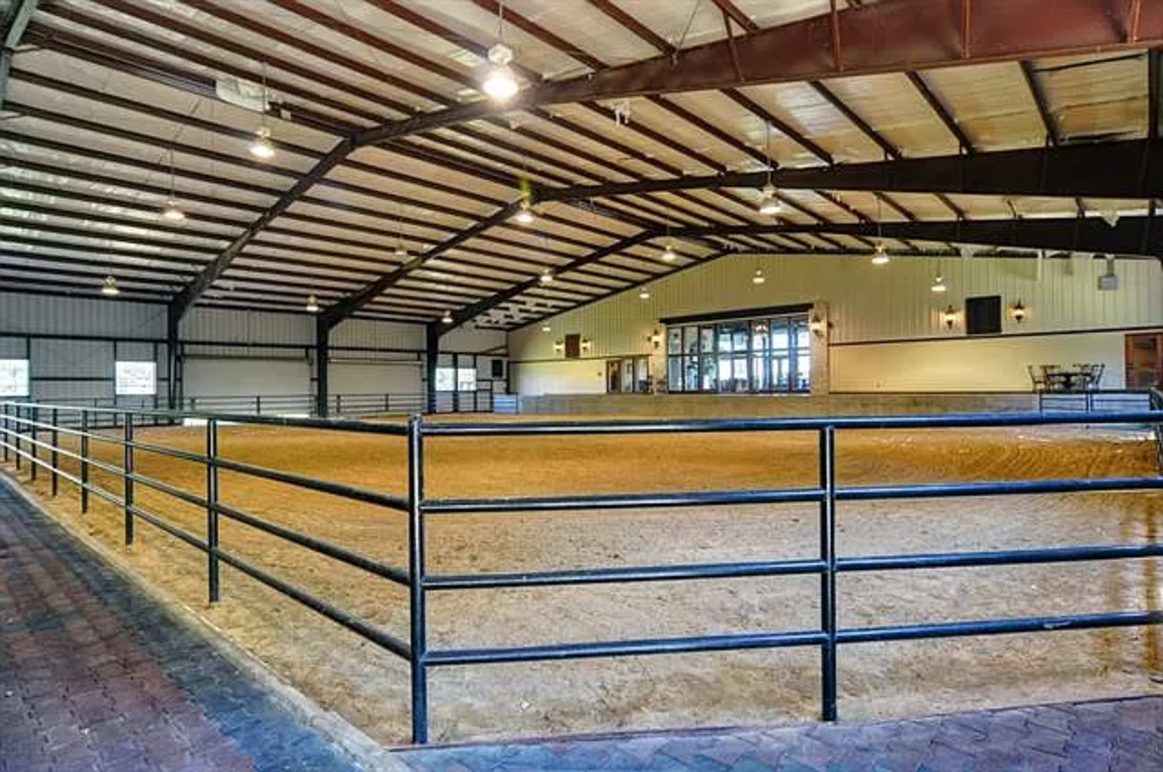 An indoor riding arena with a sandy floor, surrounded by black metal fencing and a spacious area for spectators.