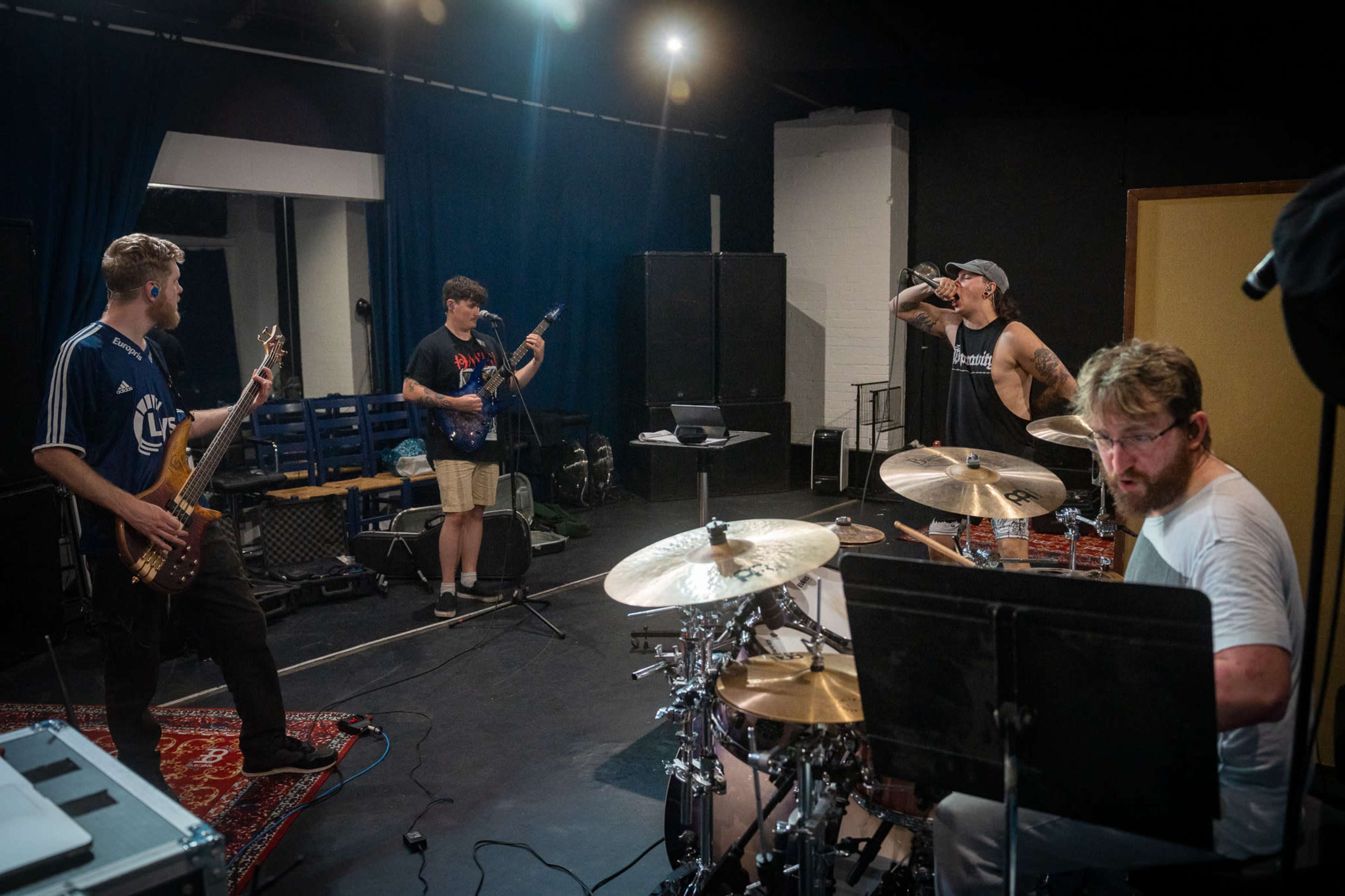 A band rehearses in a practice studio, with musicians playing electric guitar, bass, and drums while a vocalist sings.