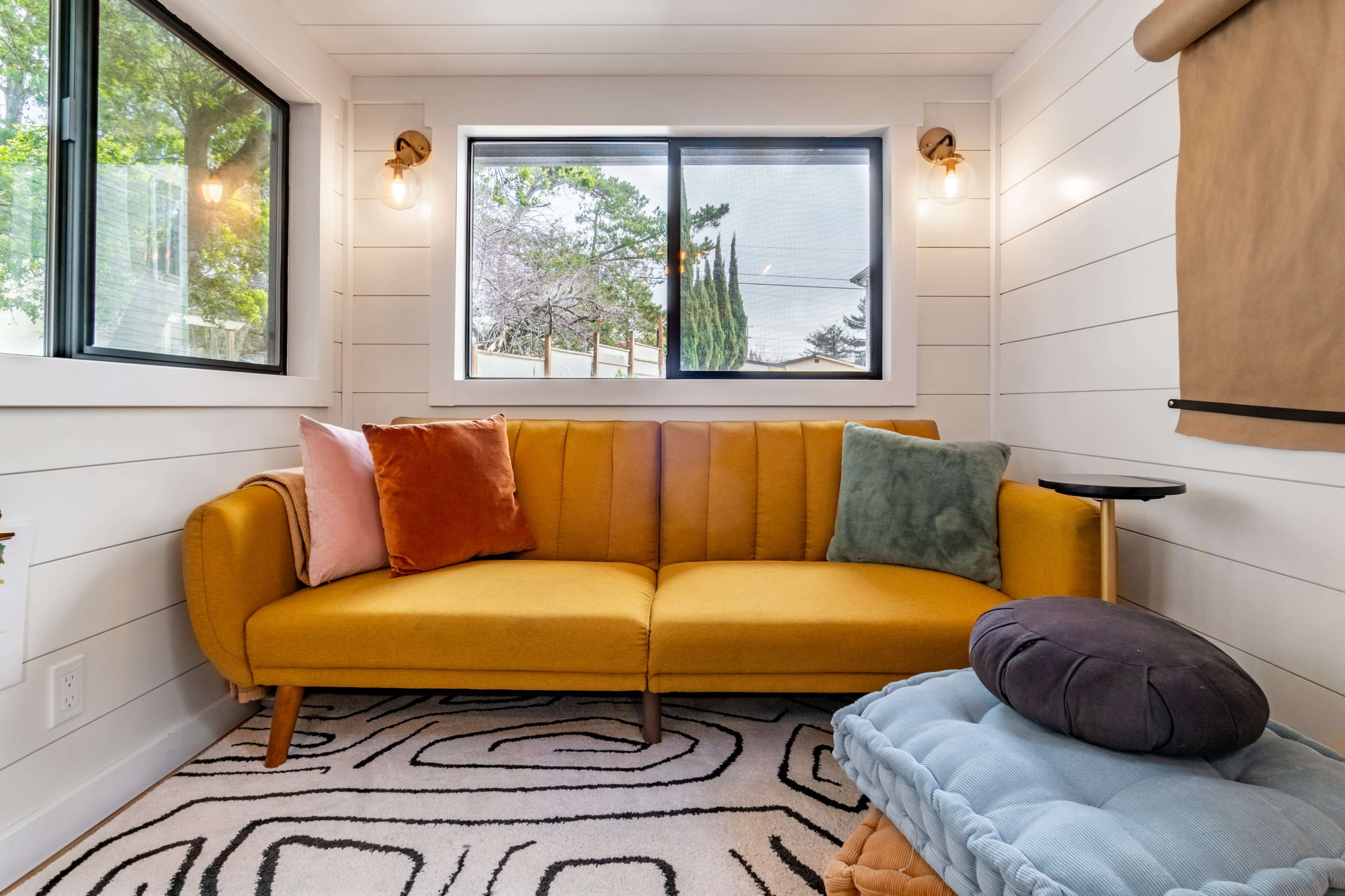 A cozy living space featuring a yellow sofa with colorful pillows, large windows allowing natural light, and a patterned rug on the floor.