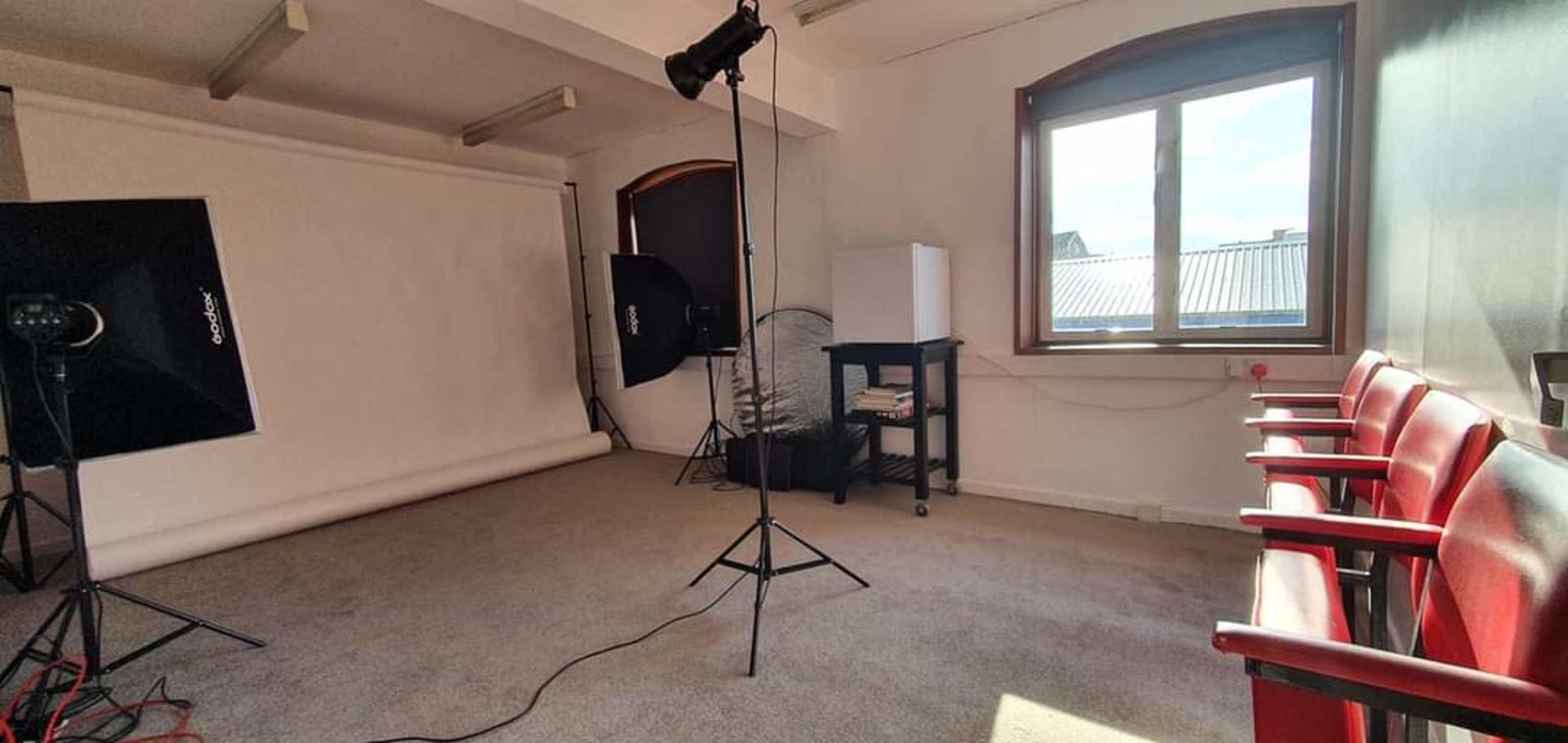 A photography studio with a backdrop, lighting equipment, a window, and several red chairs arranged against the wall.