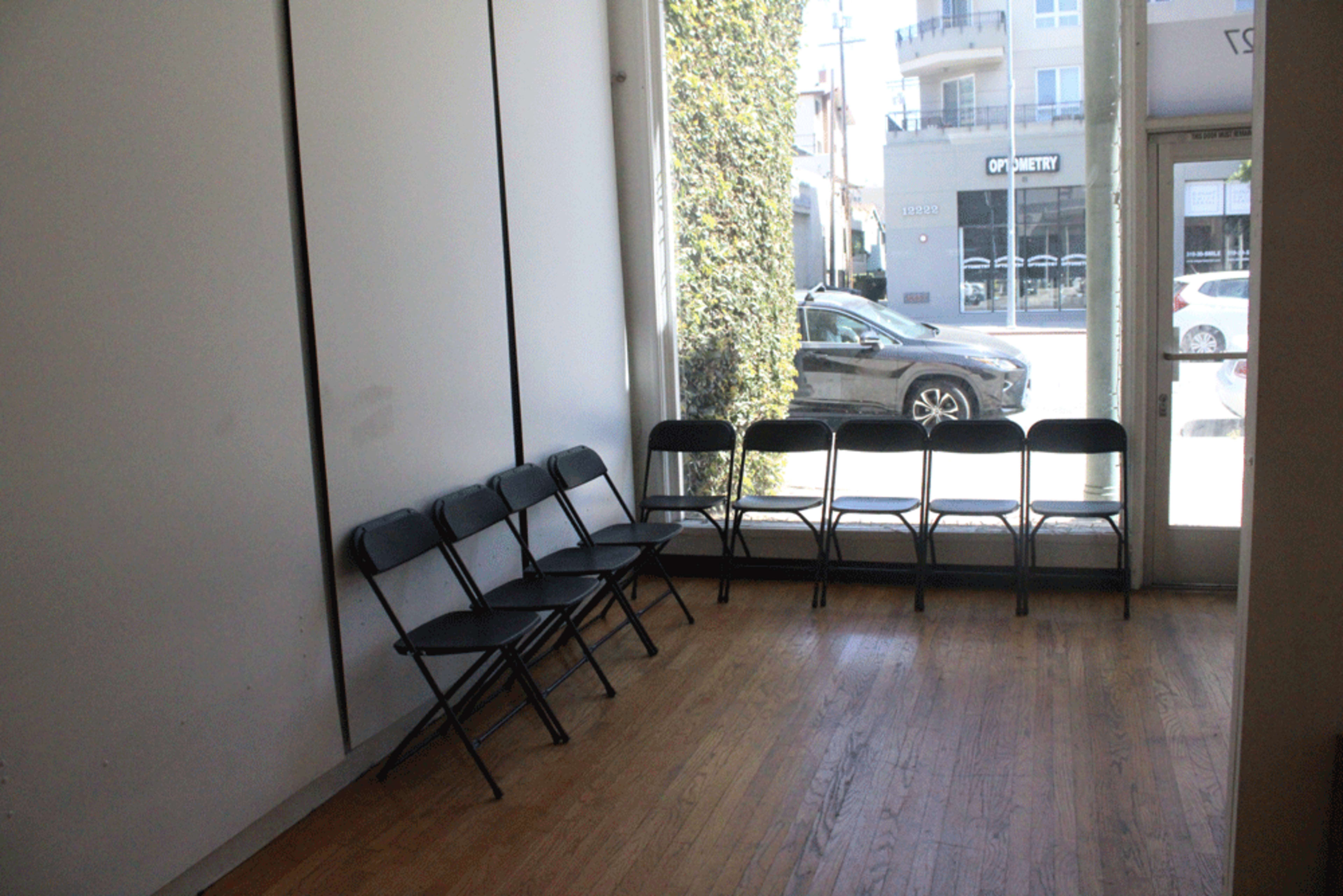 The image shows a sparse interior with a row of black folding chairs positioned against a white wall, and a view of a street and parked cars visible through large windows.