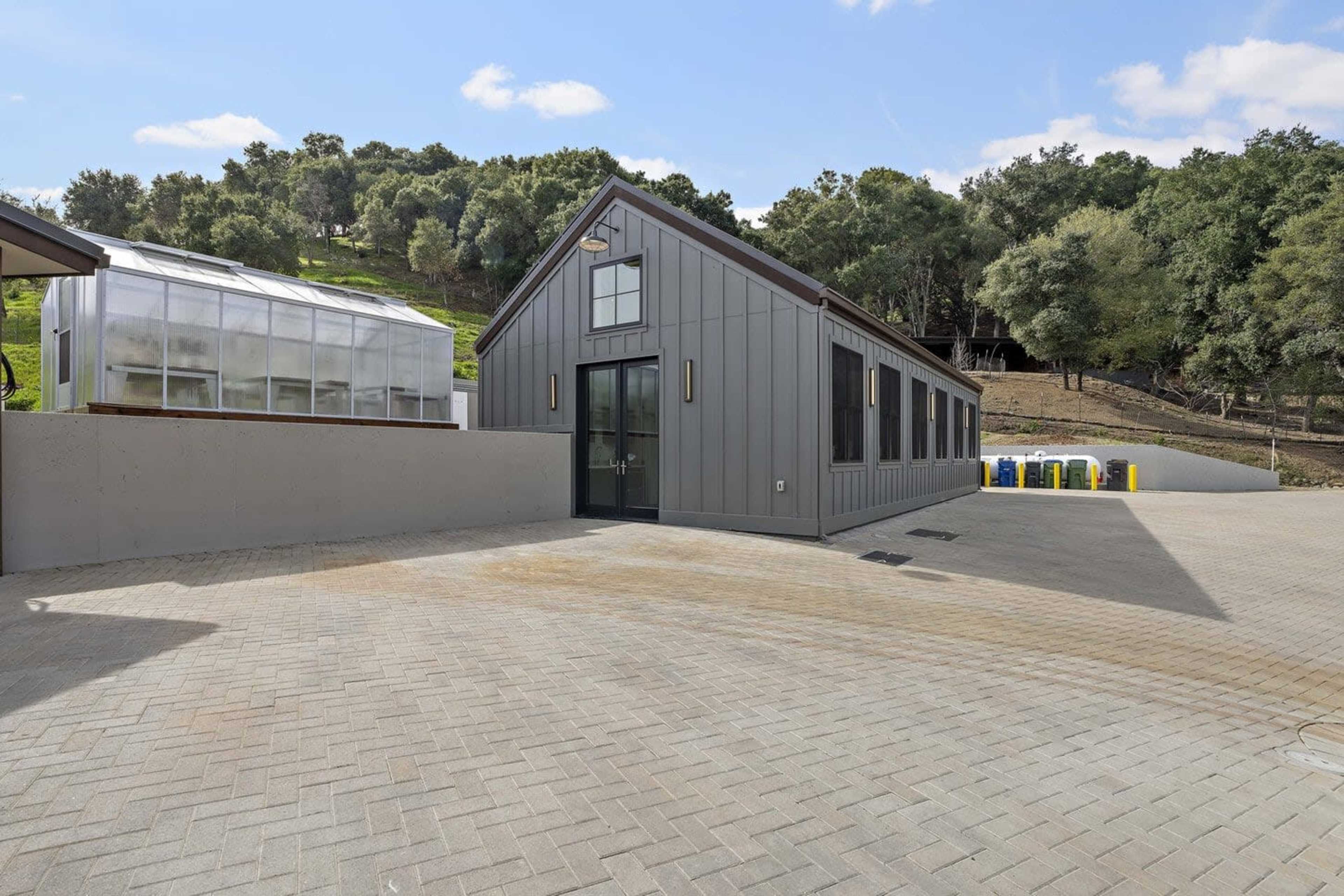 The image shows a modern, gray-clad building with a peaked roof situated next to a paved area that includes a greenhouse and outdoor facilities.