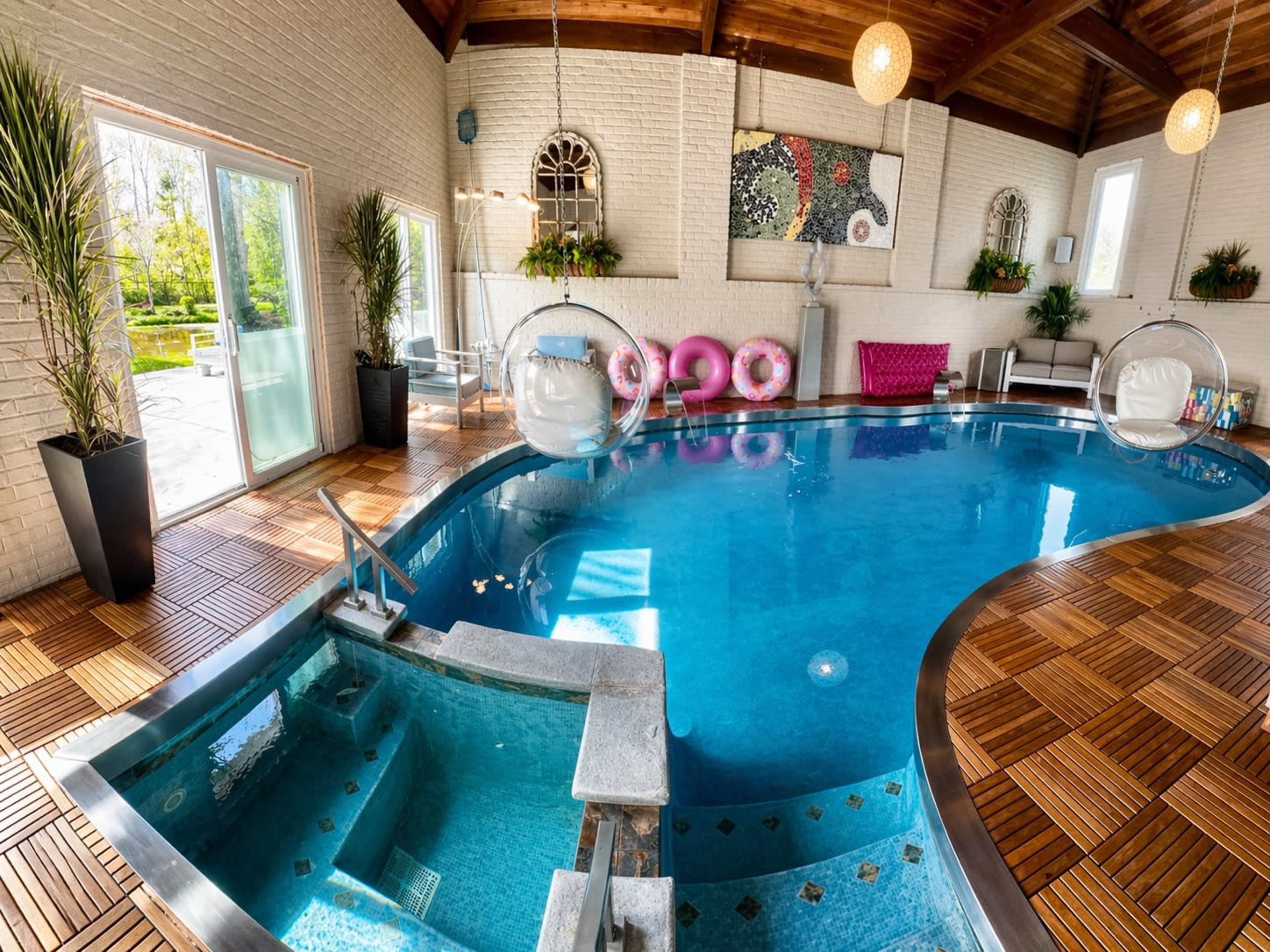 The image shows an indoor swimming pool with a hot tub, surrounded by lounge chairs and colorful floating donuts, set under a wooden ceiling with large windows.