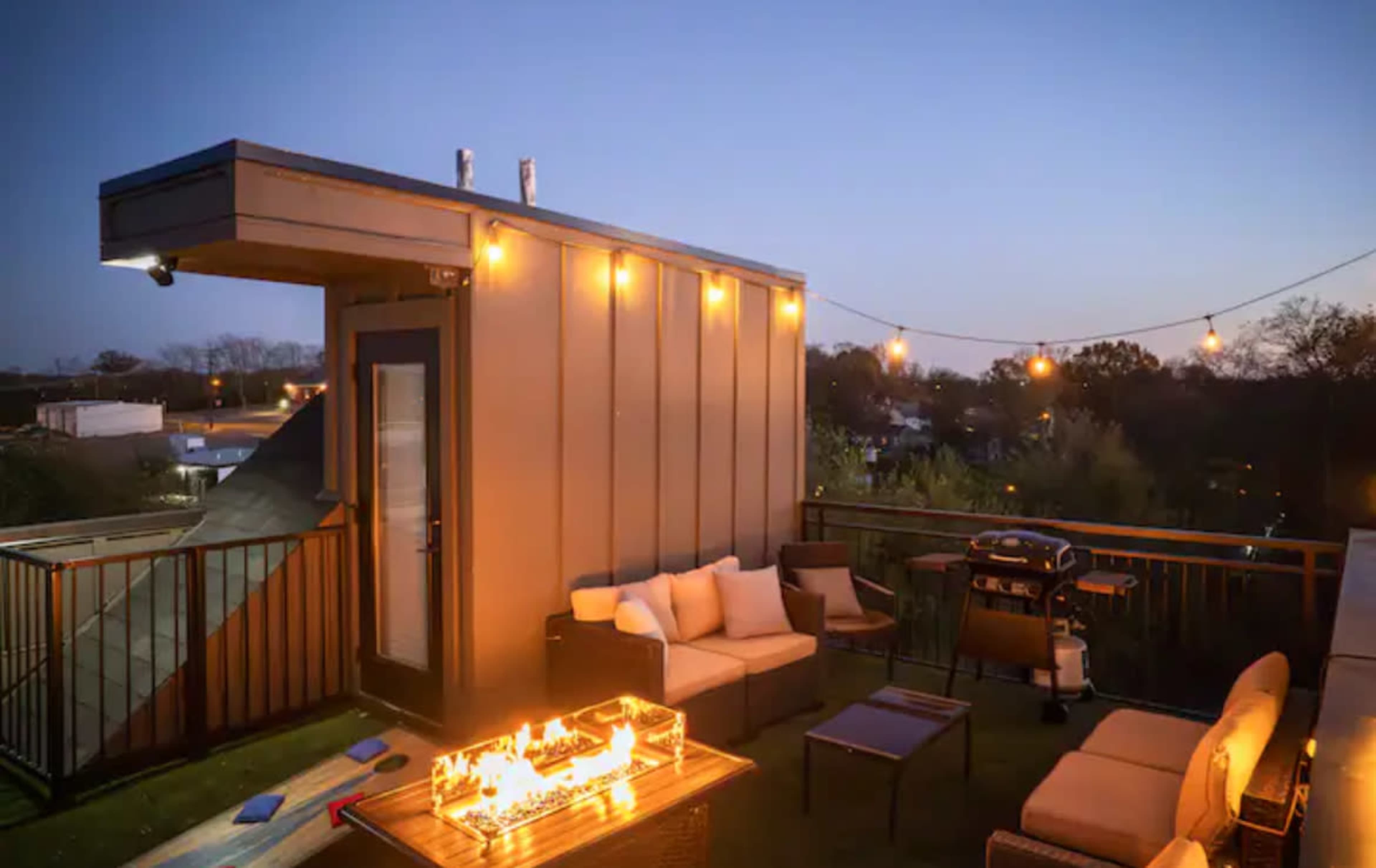 A rooftop patio features a seating area with a fire pit and a grill, illuminated by string lights during dusk.