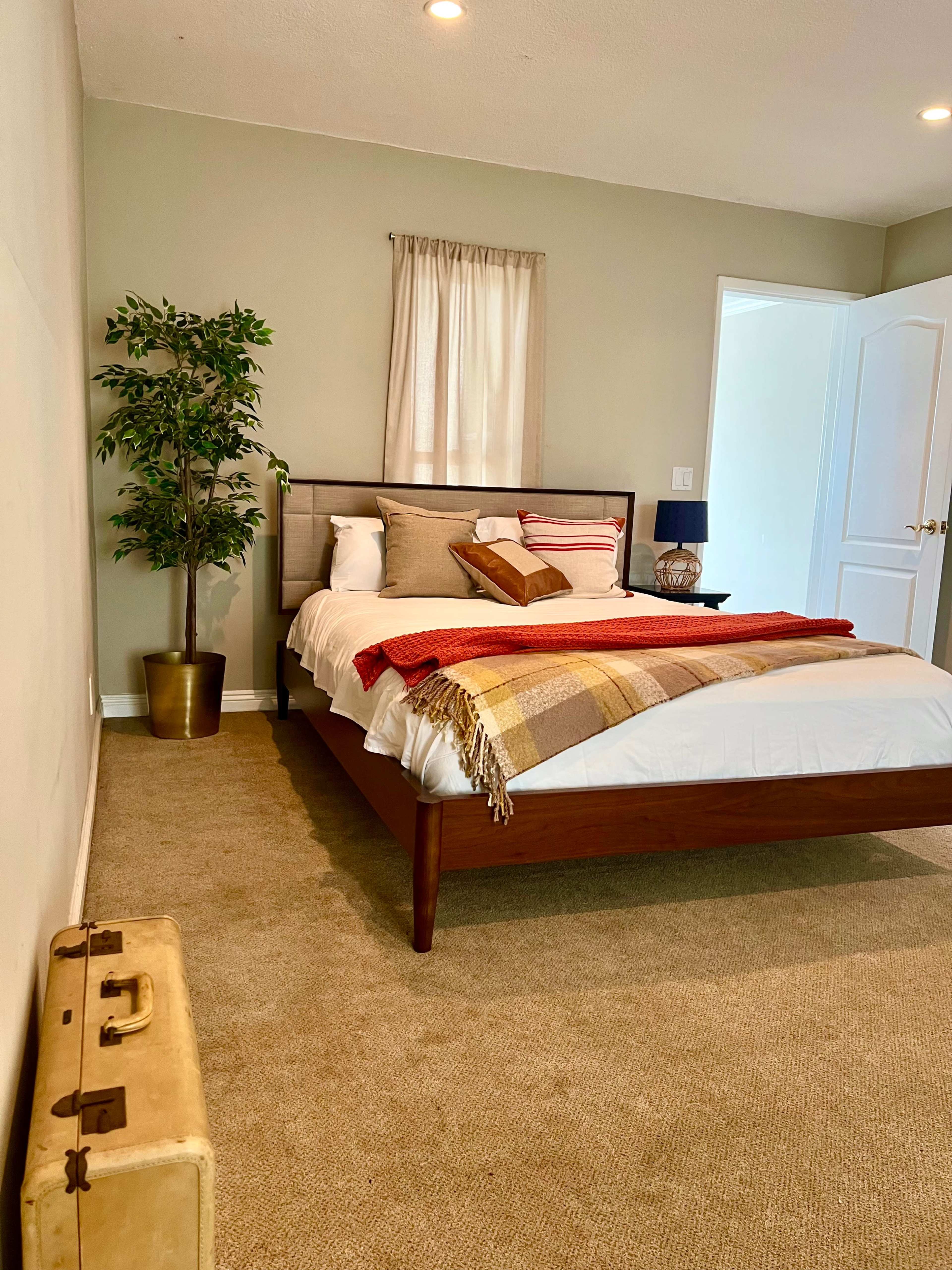 A neatly arranged bedroom featuring a wooden bed with an arrangement of pillows, a side table with a lamp, and a suitcase positioned on the carpeted floor.