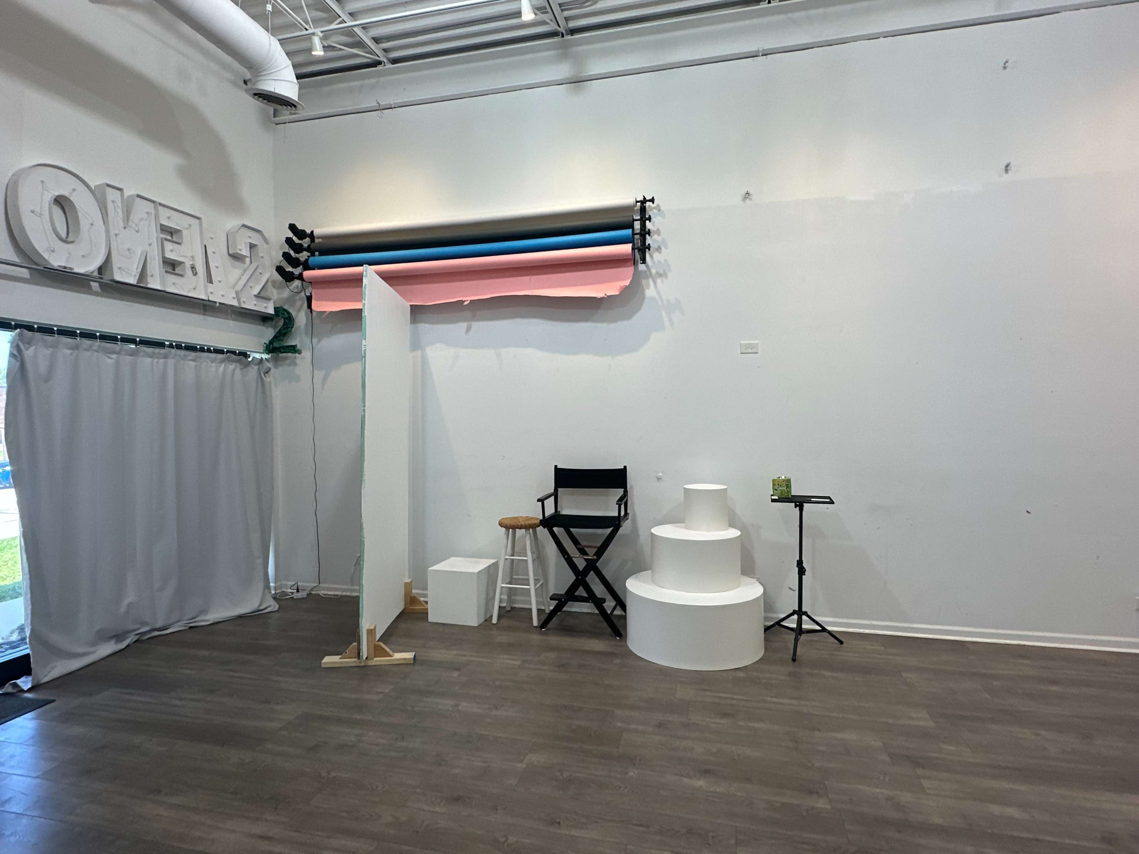 The image shows a minimalist studio space featuring a backdrop of colored paper rolls, a stool, and three white circular platforms against a plain wall.