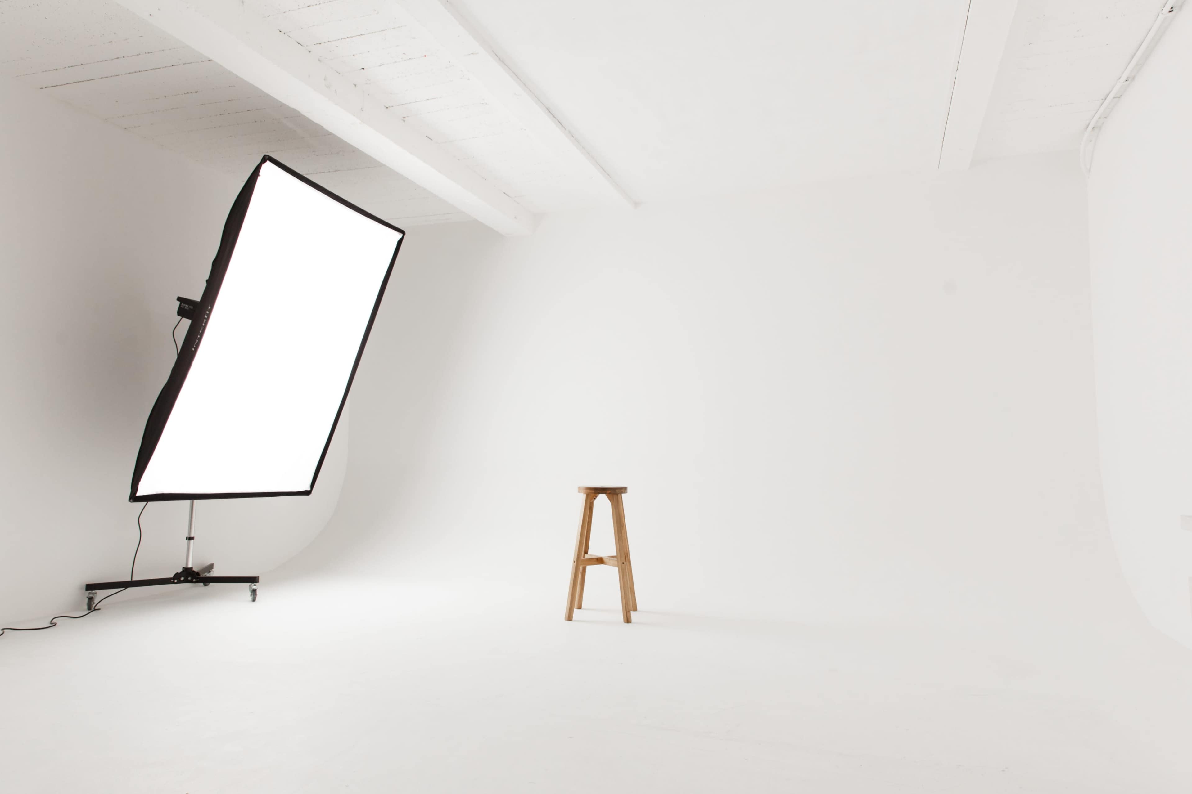 A minimalist photography studio featuring a white backdrop, a light source on a stand, and a wooden stool positioned in the center.