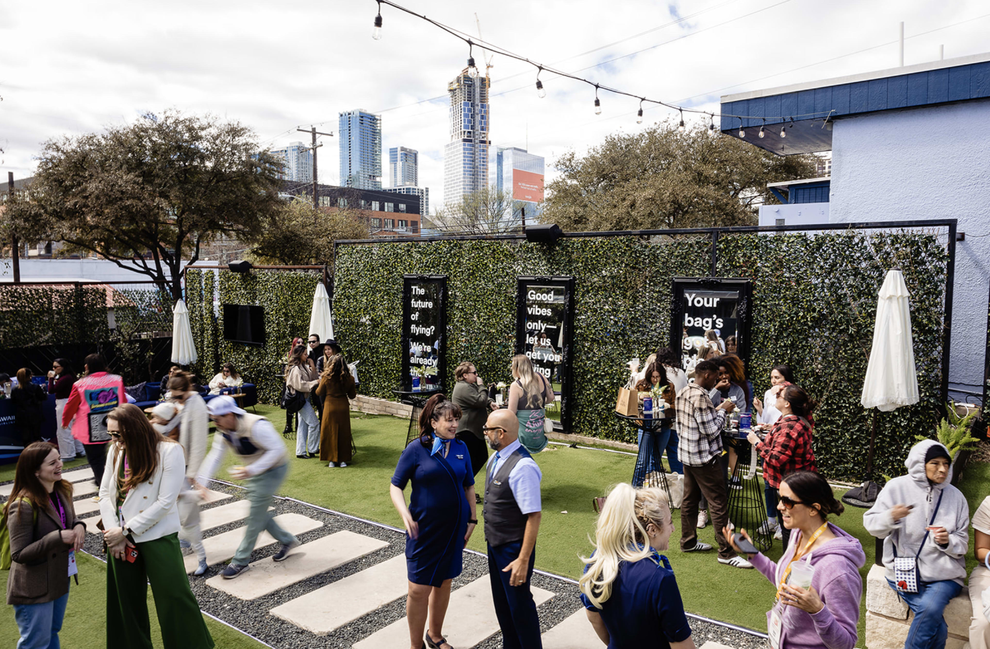 A group of people socialize and mingle in a lively outdoor space adorned with greenery and string lights, while city buildings loom in the background.