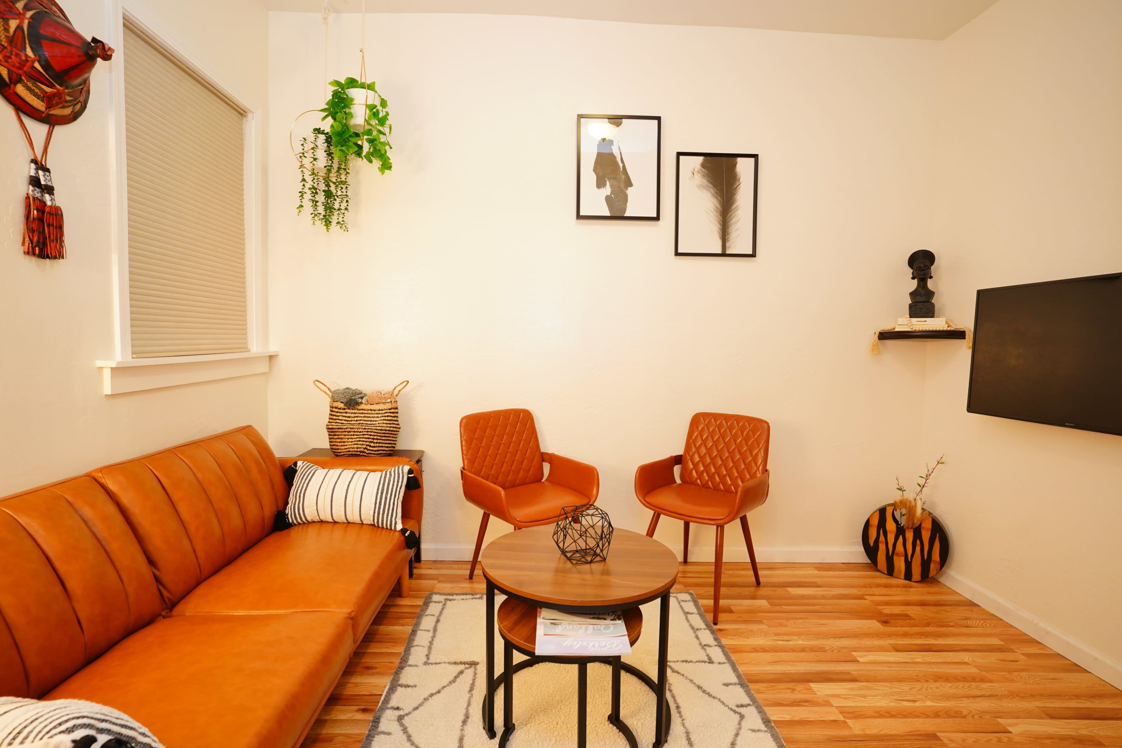 The image shows a cozy living room featuring a brown leather sofa, two orange armchairs, a round coffee table, and decorative wall art.