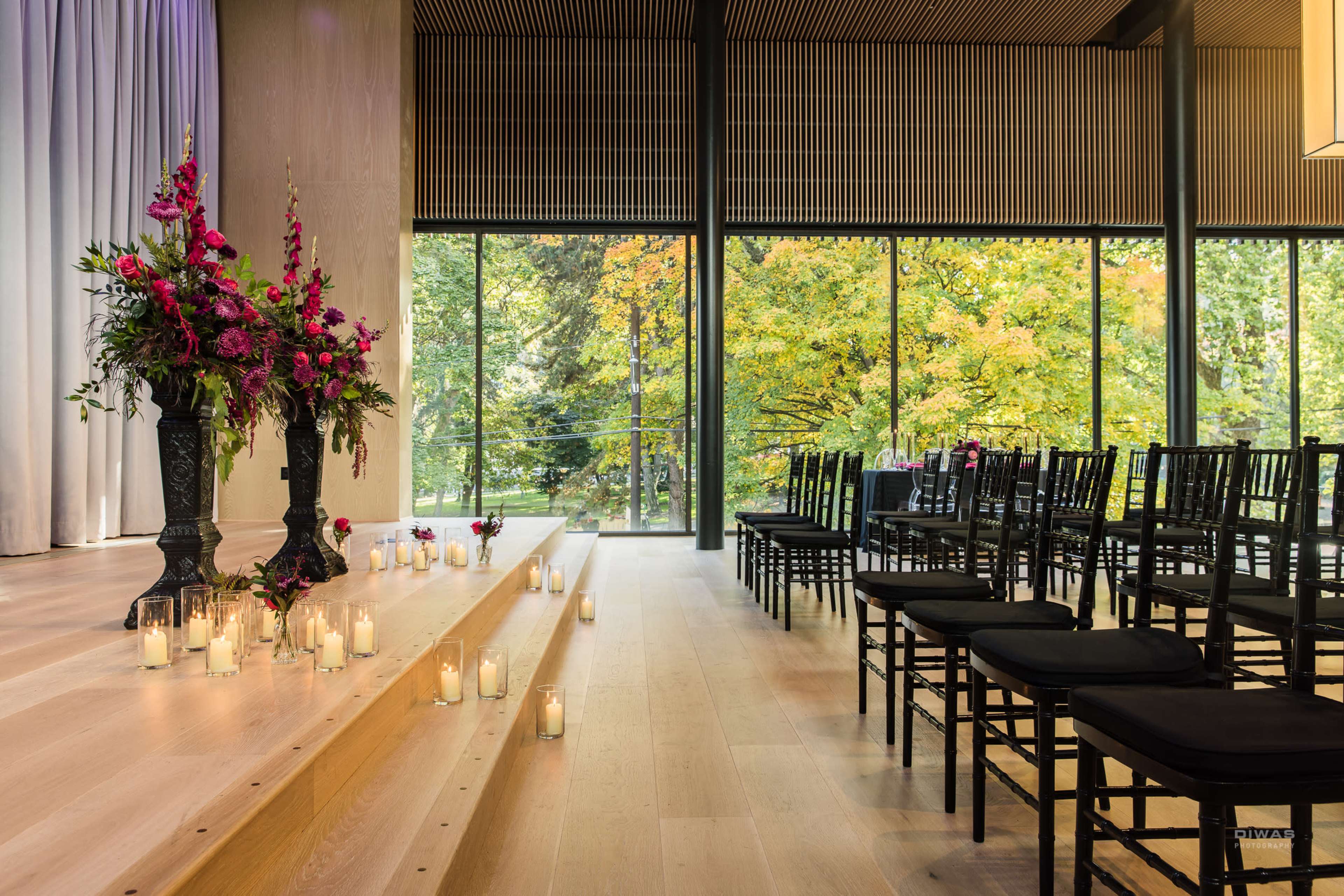 A wedding ceremony setup features a floral arrangement on a stage with candles and rows of black chairs facing a large window overlooking autumn trees.