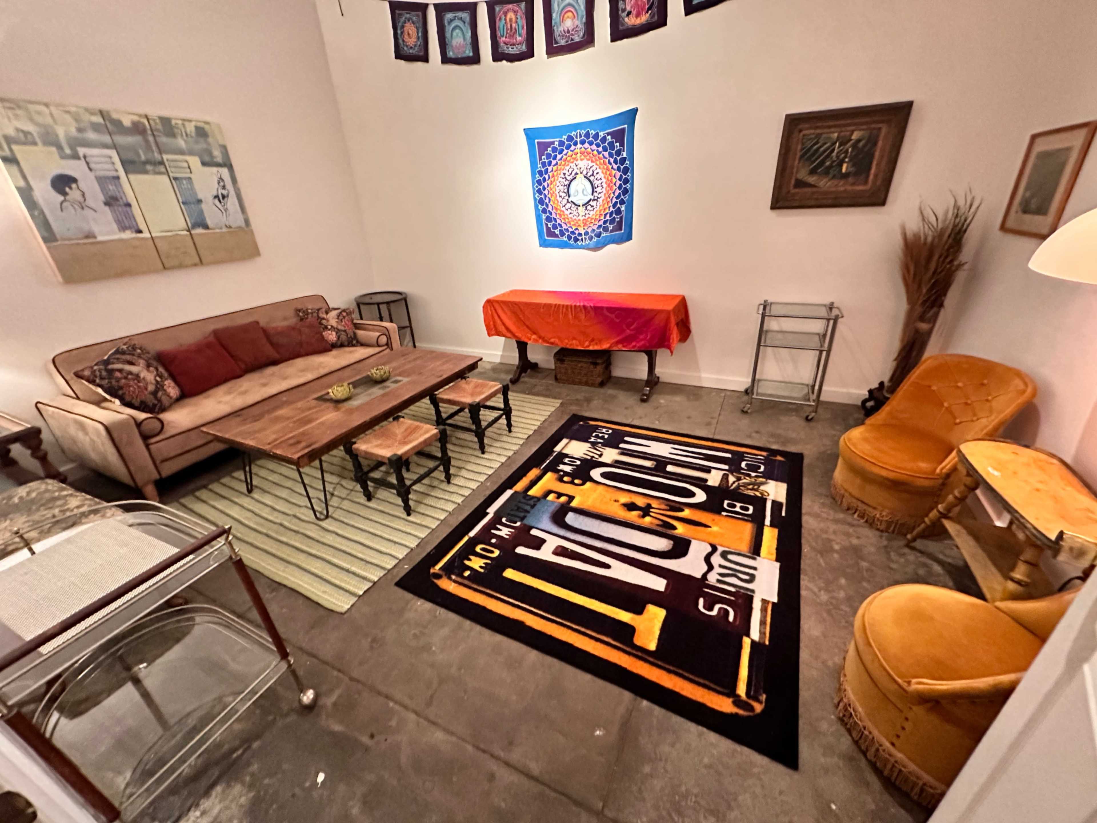 The image shows a cozy room featuring a red sofa, a wooden coffee table with decorative items, an orange tablecloth-covered table, and two yellow chairs, all set against a backdrop of wall art and a patterned rug.