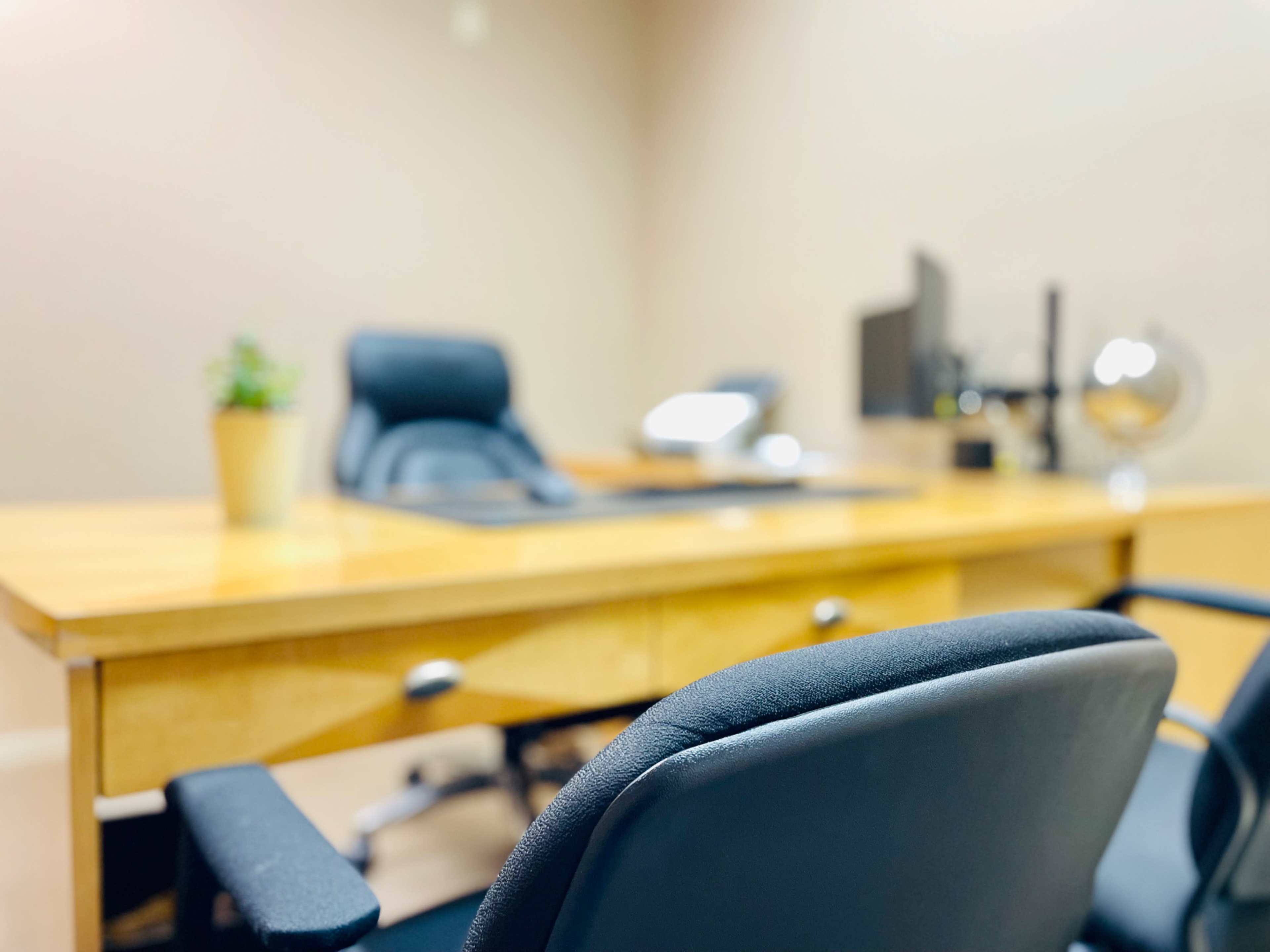 The image shows a modern office with a wooden desk, black chairs, and a small potted plant.
