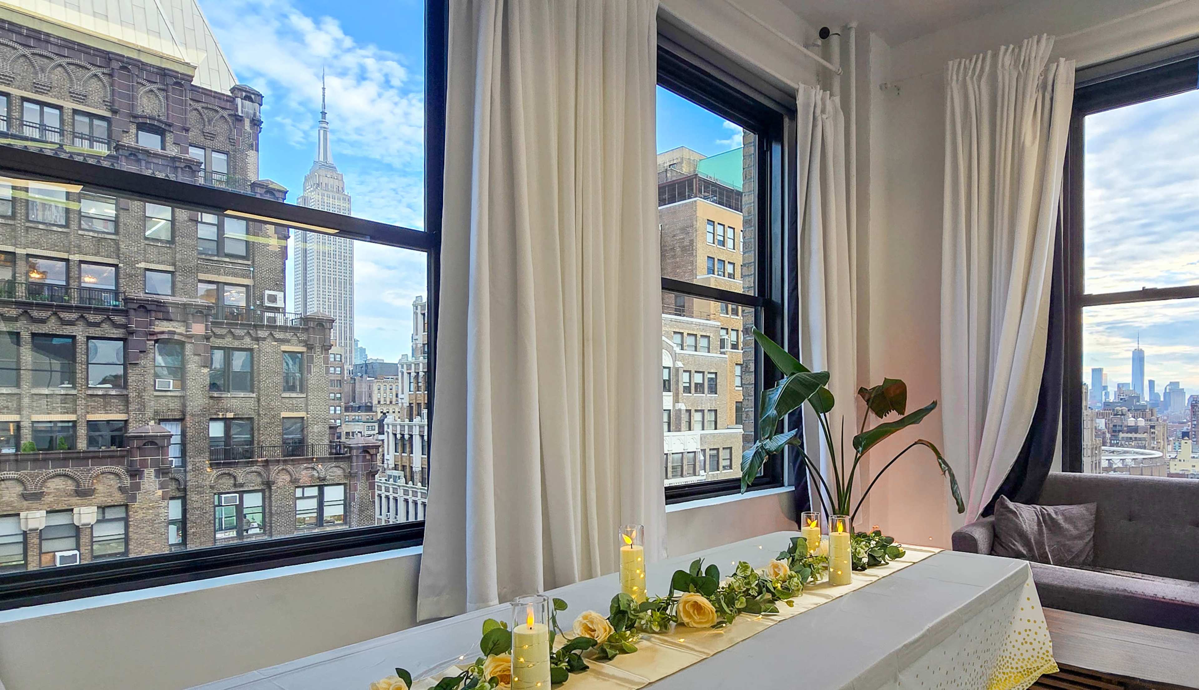 The scene features a table set with candles and greenery near large windows that overlook a cityscape, including the Empire State Building.