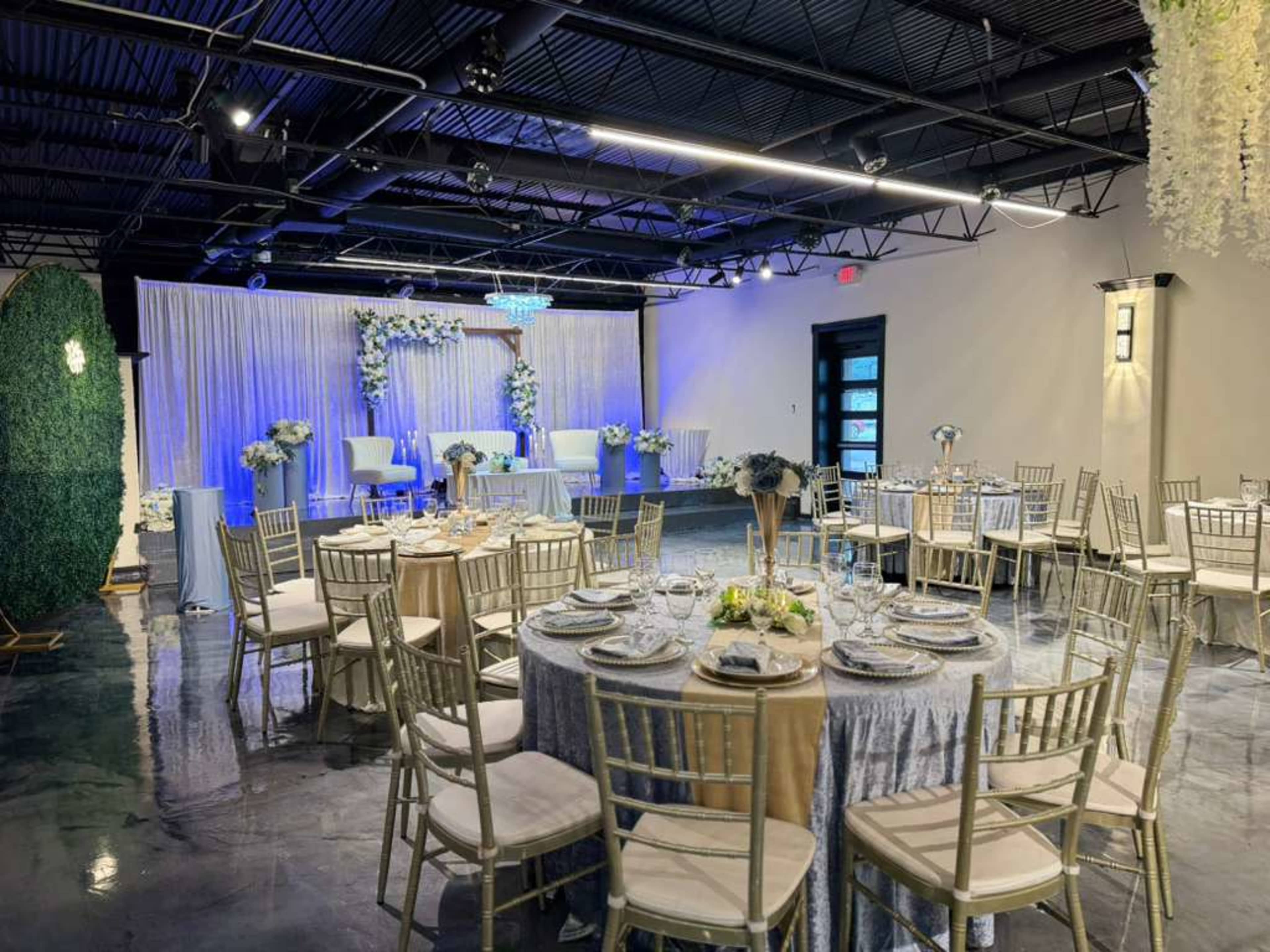 The image shows a decorated event space with round tables set for a gathering, featuring floral centerpieces and gold chairs, and a backdrop of draped fabric and decorative elements at one end.