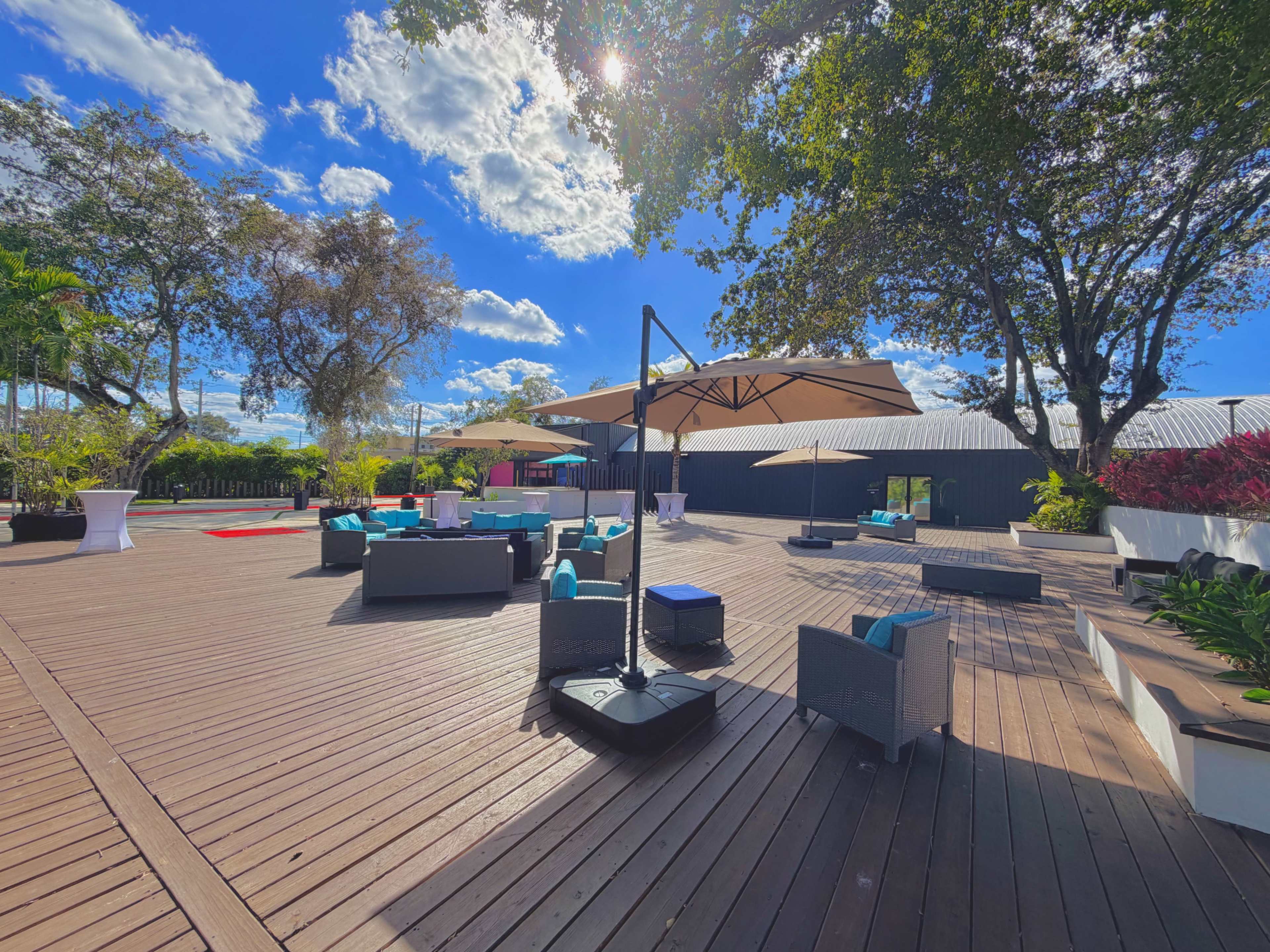 The image shows a spacious outdoor deck area with modern furniture, umbrellas, and green landscaping under a bright blue sky.