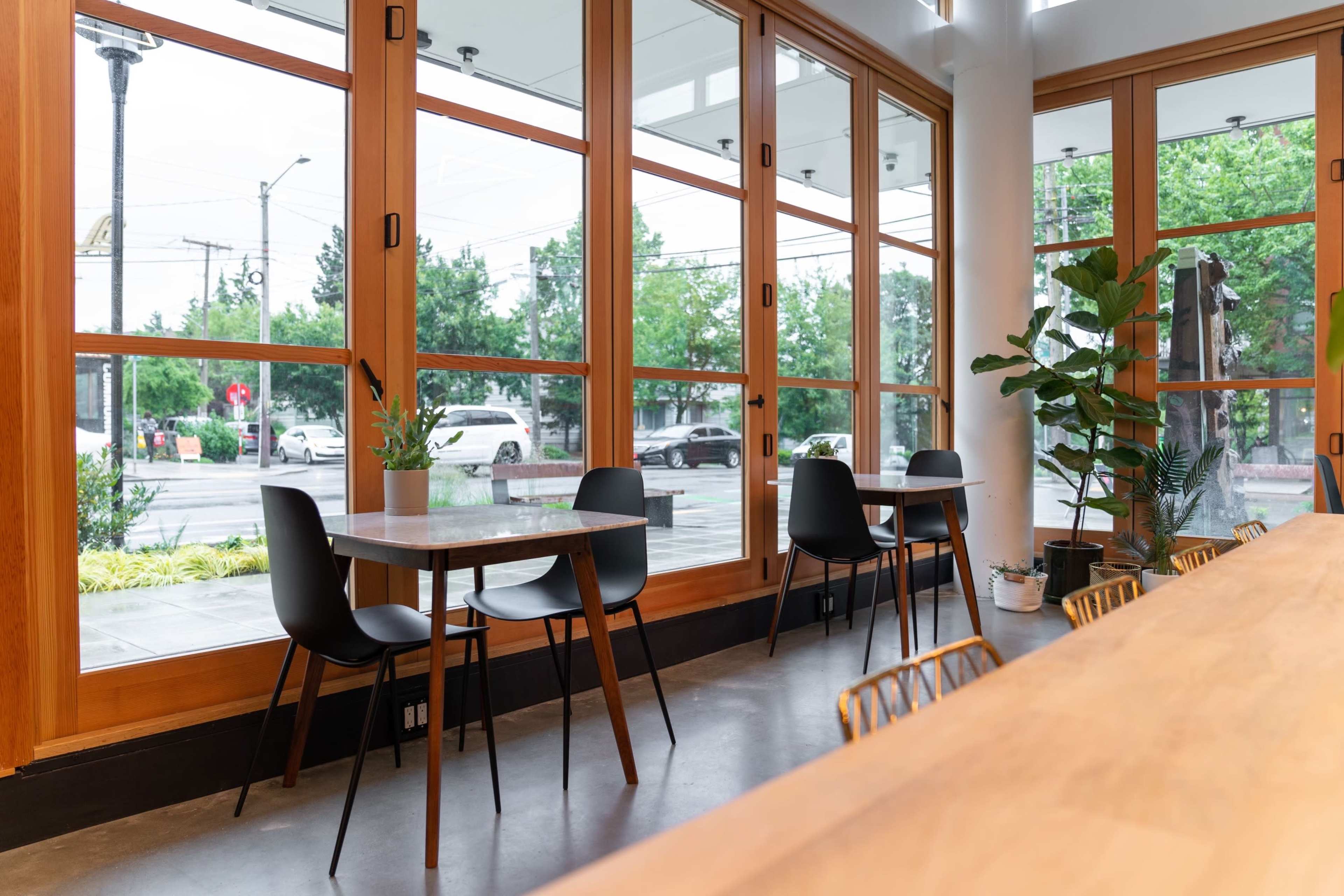 A modern café interior with wooden tables and black chairs beside large windows, offering a view of the outside street and greenery.