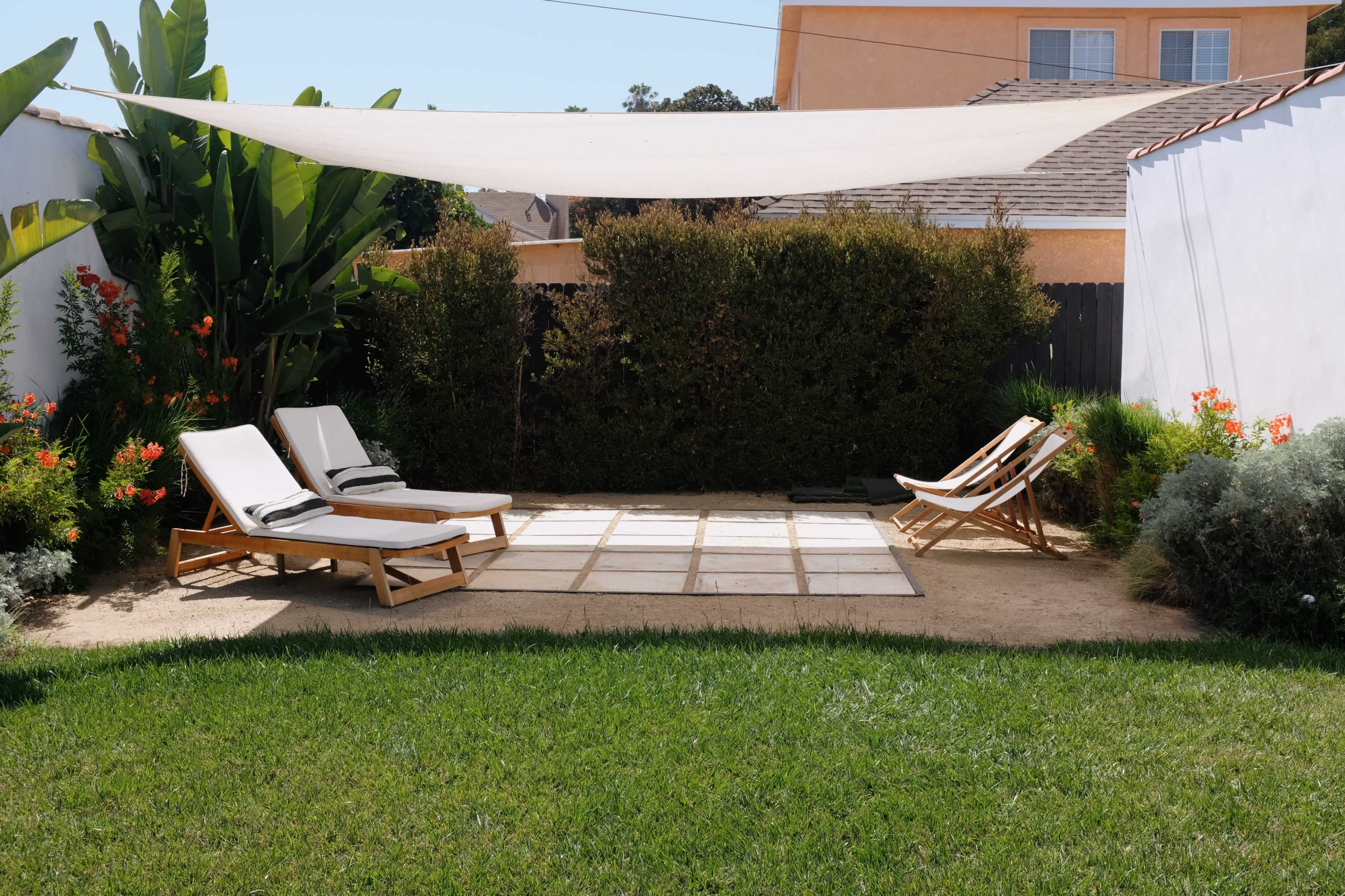 The image shows a landscaped backyard with two lounge chairs and a seating area under a fabric sunshade, surrounded by greenery and flowering plants.