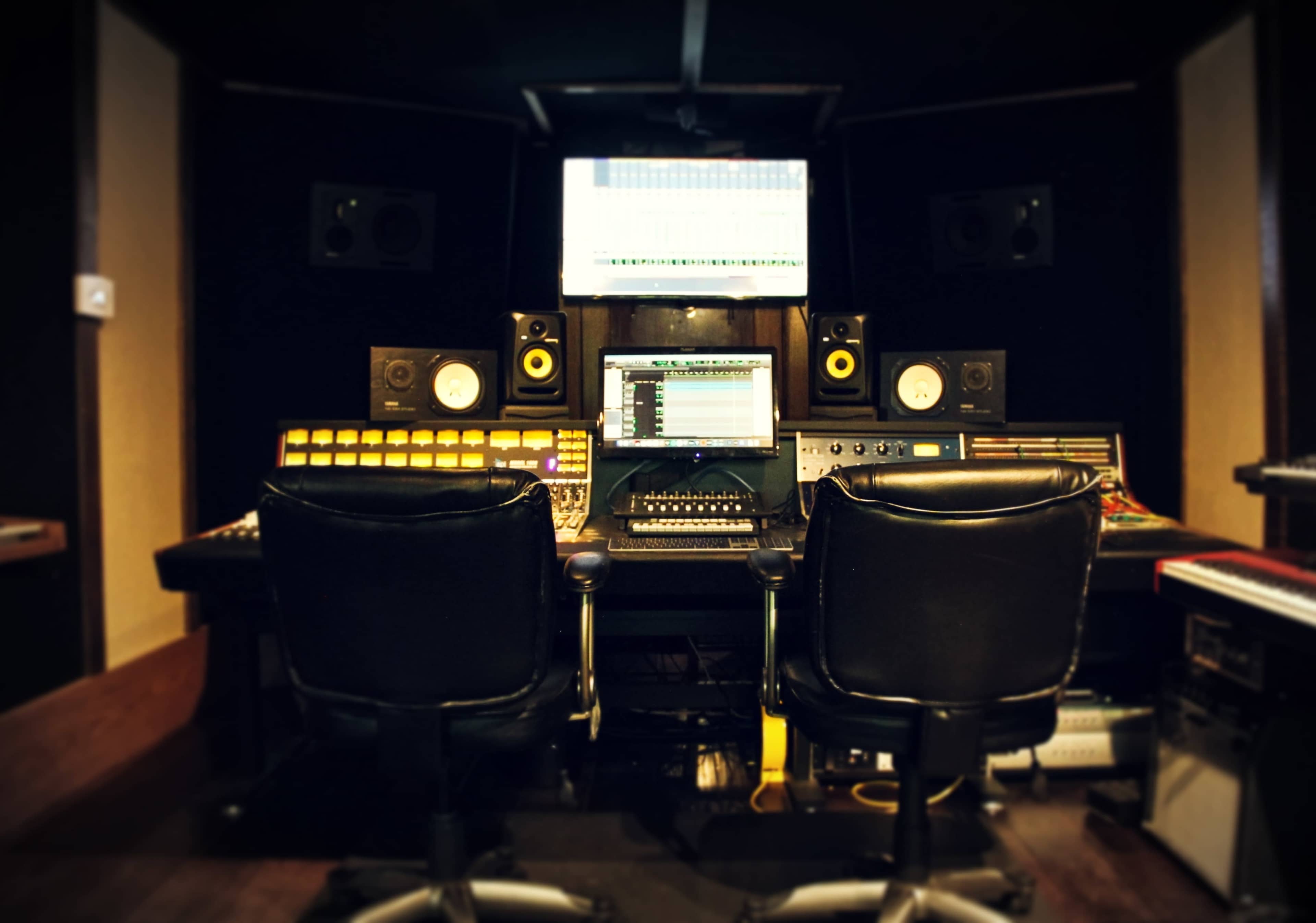 A music production studio featuring a control desk with multiple sliders, knobs, and screens, along with two black office chairs positioned in front.