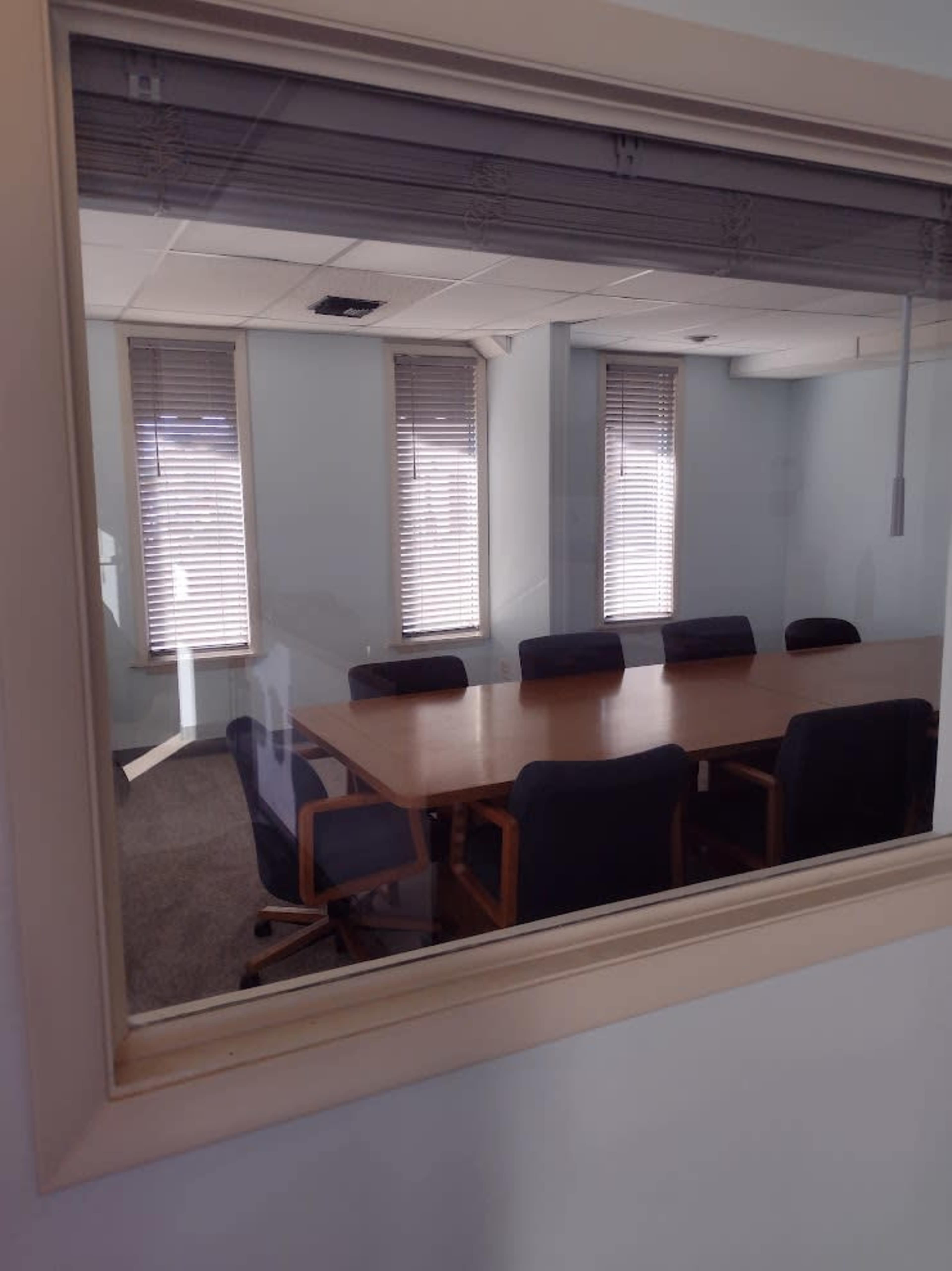 The image shows a conference room with a large table surrounded by several chairs, viewed through a glass window.