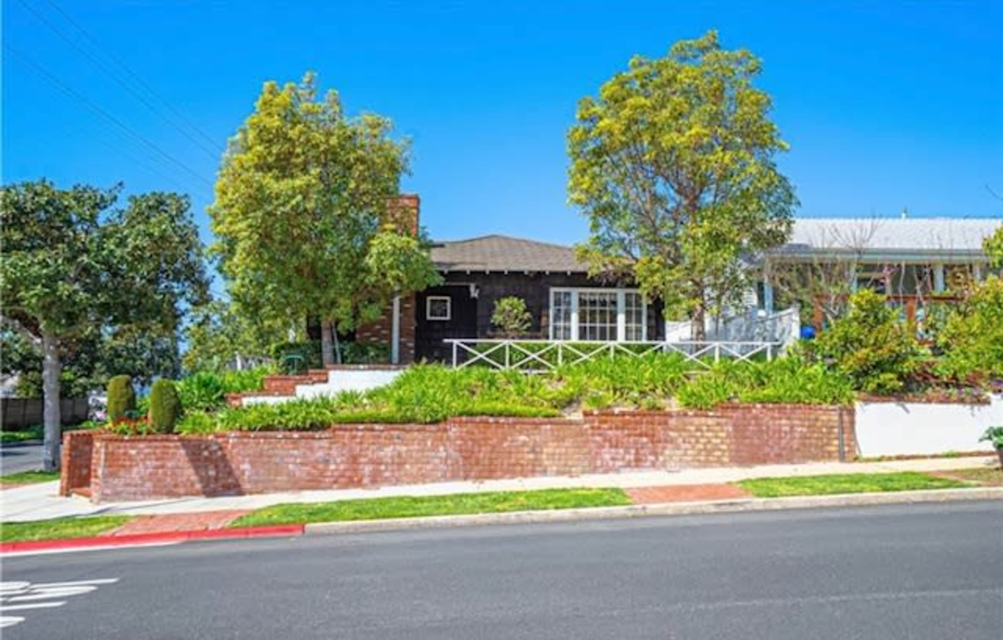 The image shows a residential street view of a single-story house with a front yard lush with greenery and a brick wall.