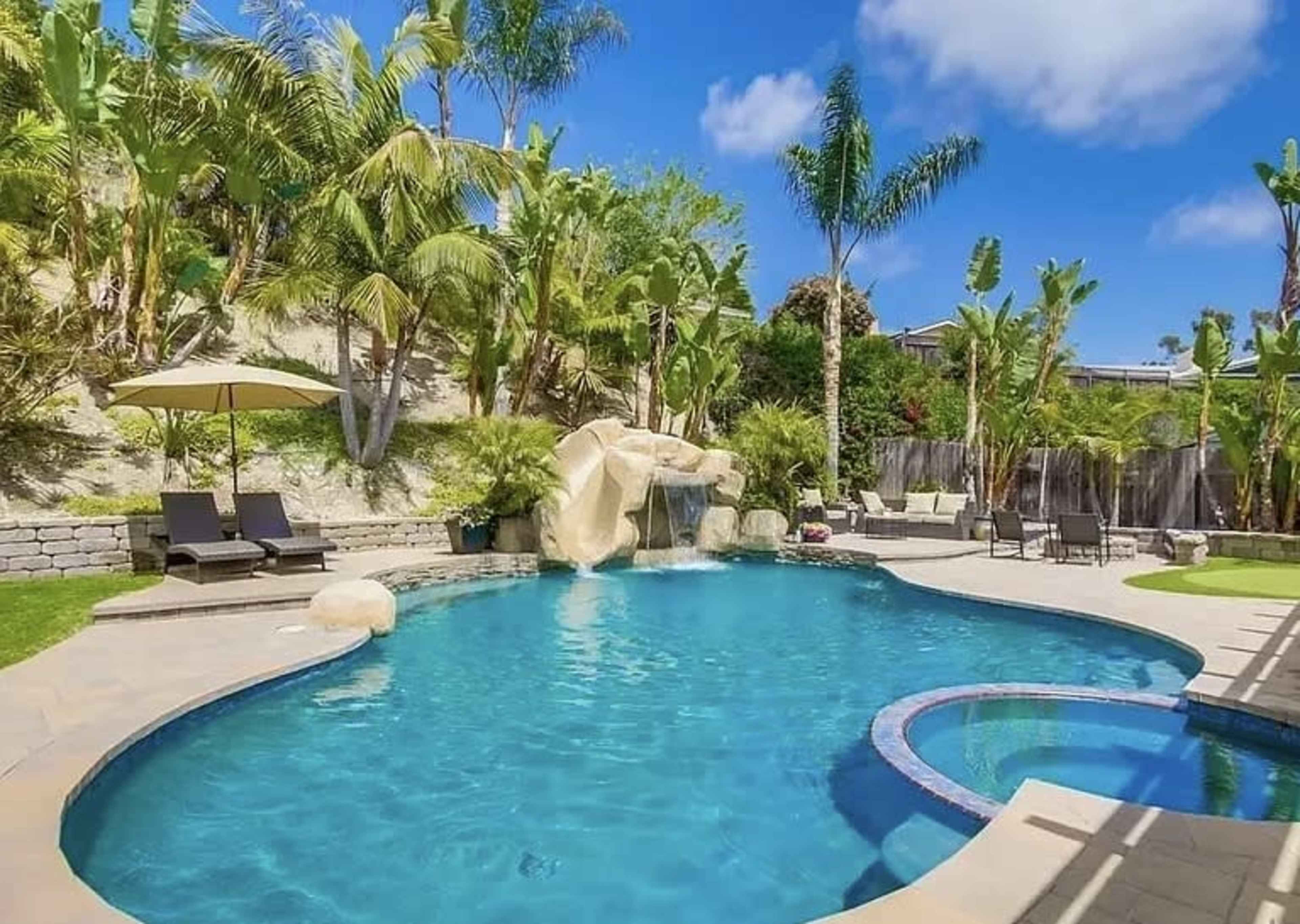 The image shows a landscaped backyard with a swimming pool featuring a rock waterfall, surrounded by tropical plants and lounge chairs under an umbrella.