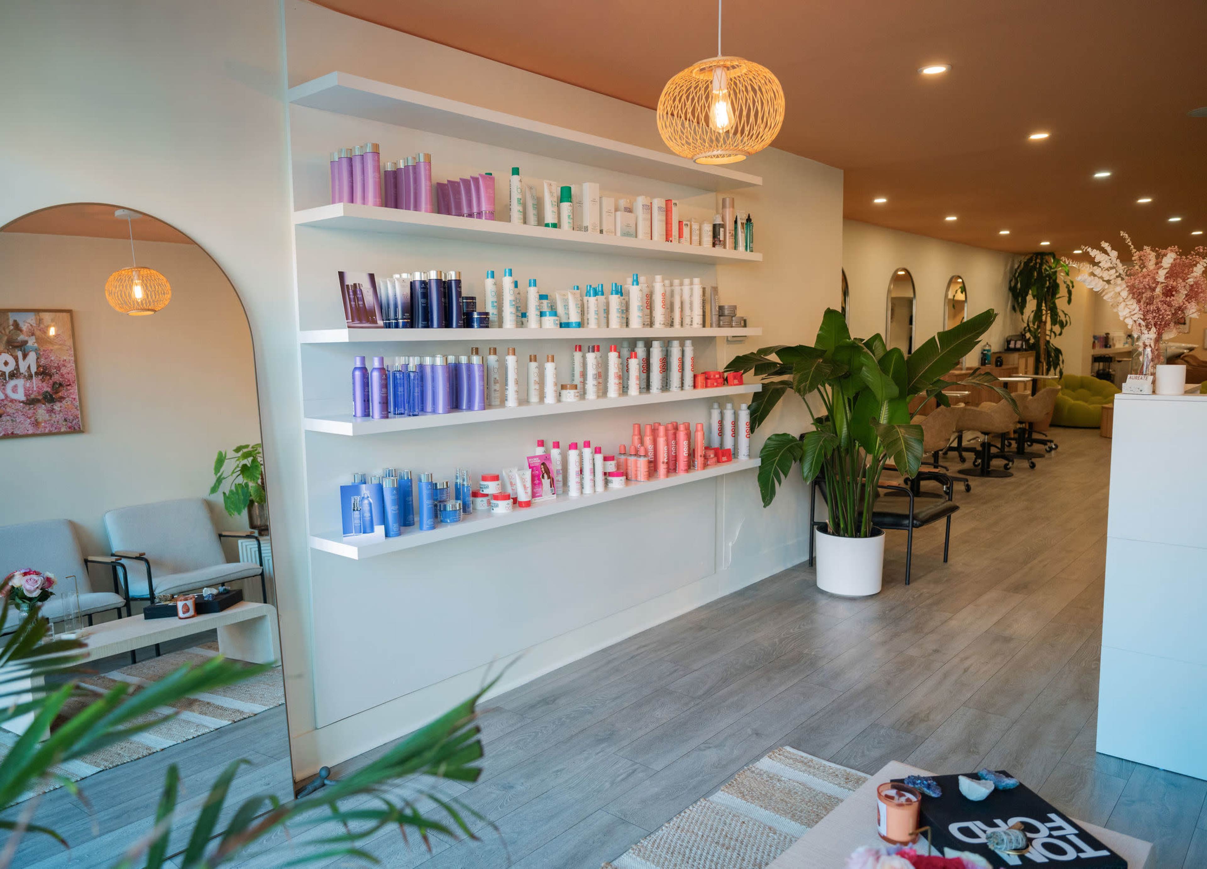The image shows a modern salon interior featuring a white wall displaying neatly arranged haircare products, with a seating area and plants in the background.