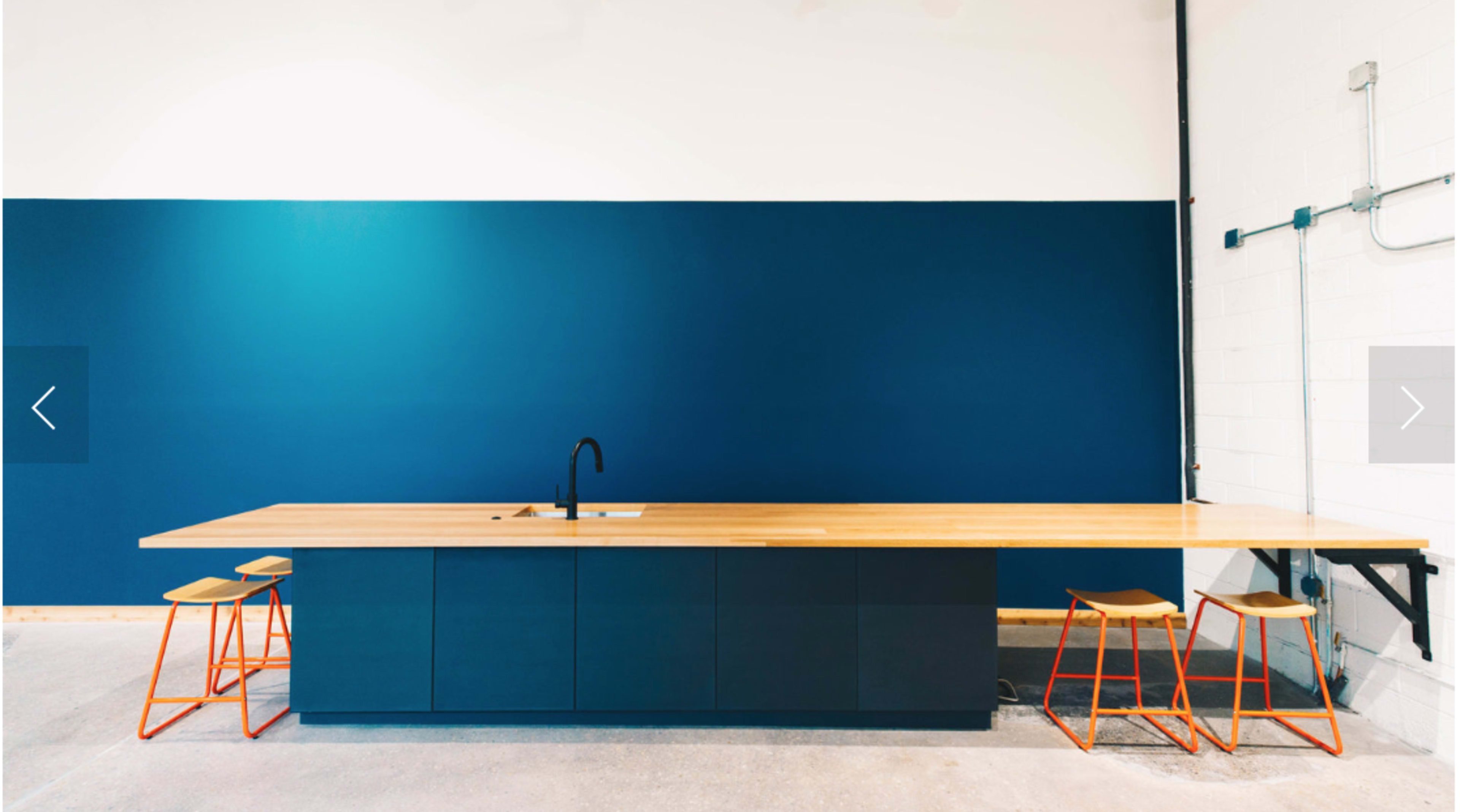 The image shows a modern kitchen featuring a long wooden countertop with a sink and dark cabinetry, complemented by three orange stools against a deep blue wall.
