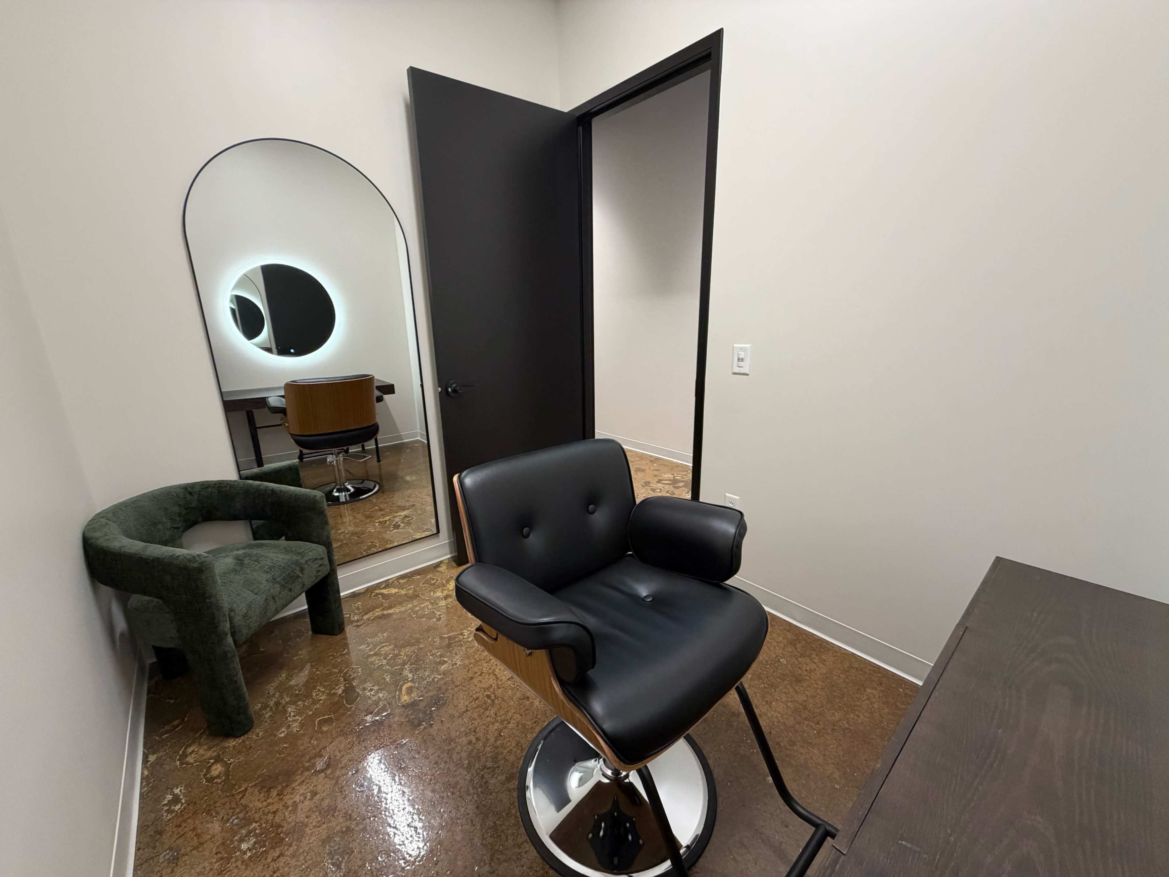 The image shows a hair salon room featuring a black salon chair, a round mirror on the wall, and a green upholstered chair in the corner.