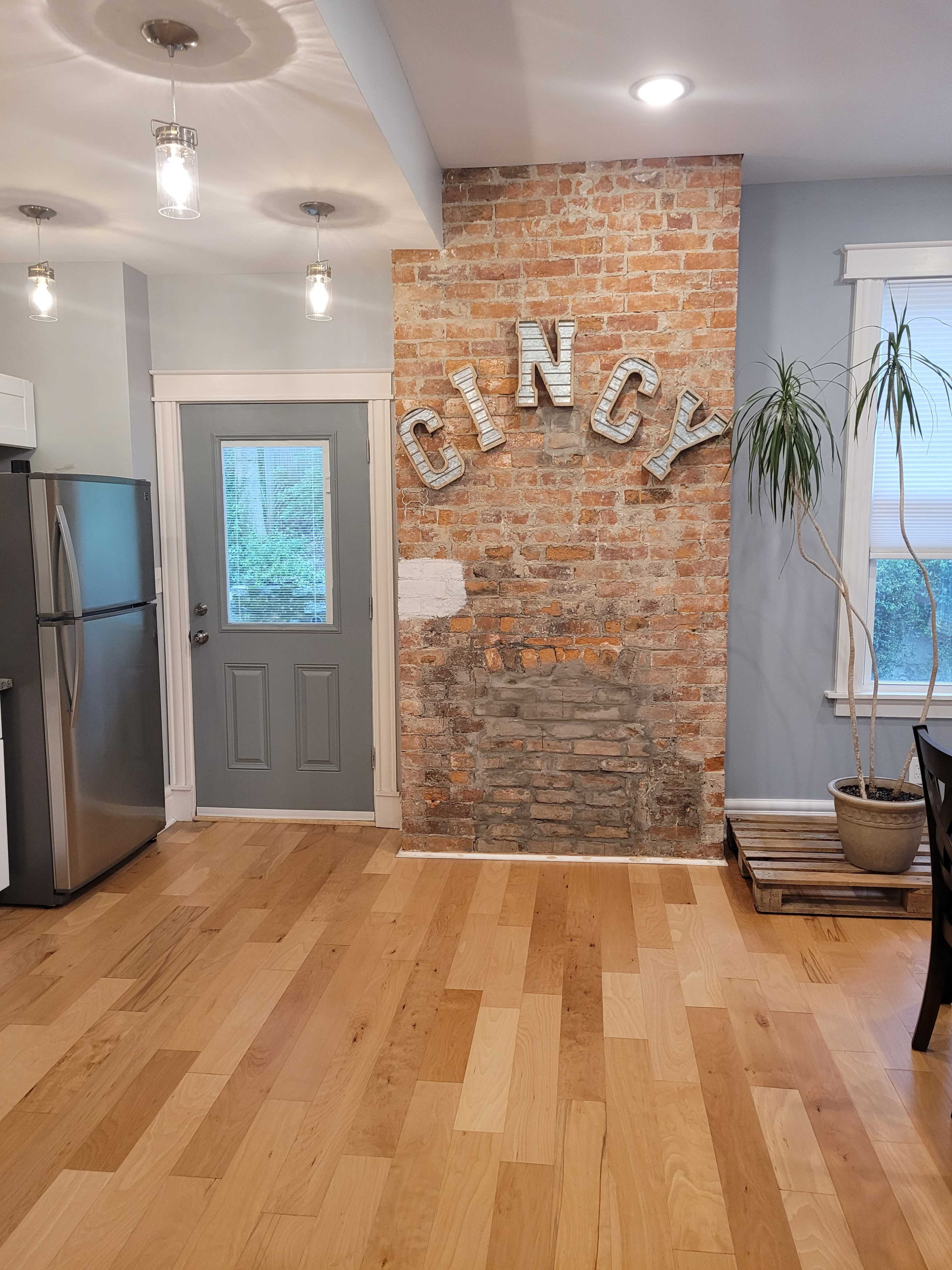 The image shows a kitchen area with a brick accent wall featuring the word "CINCY," a gray door leading outside, and a potted plant in the corner.