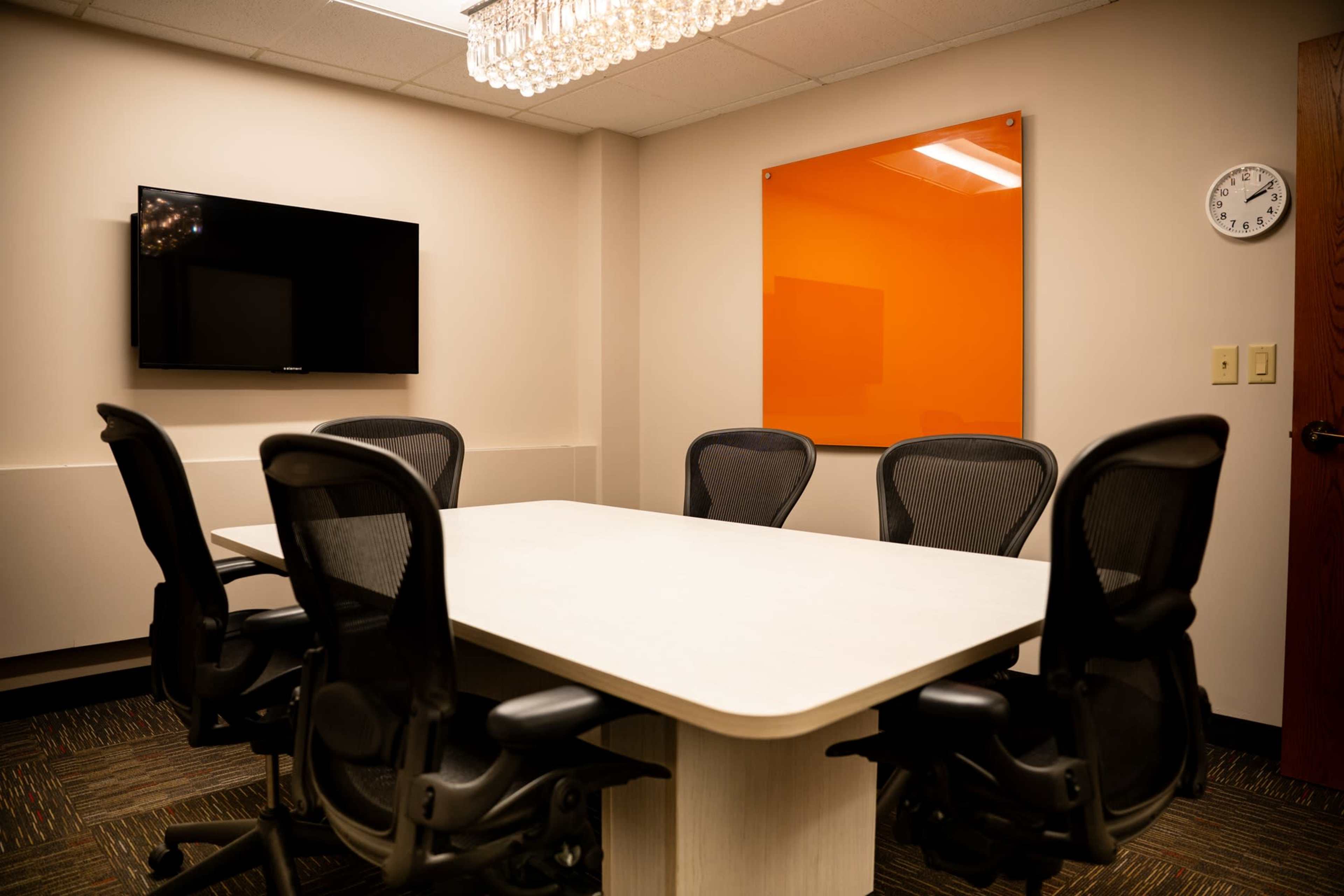 A conference room features a large table surrounded by six ergonomic chairs, a wall-mounted TV, and an orange glass panel.